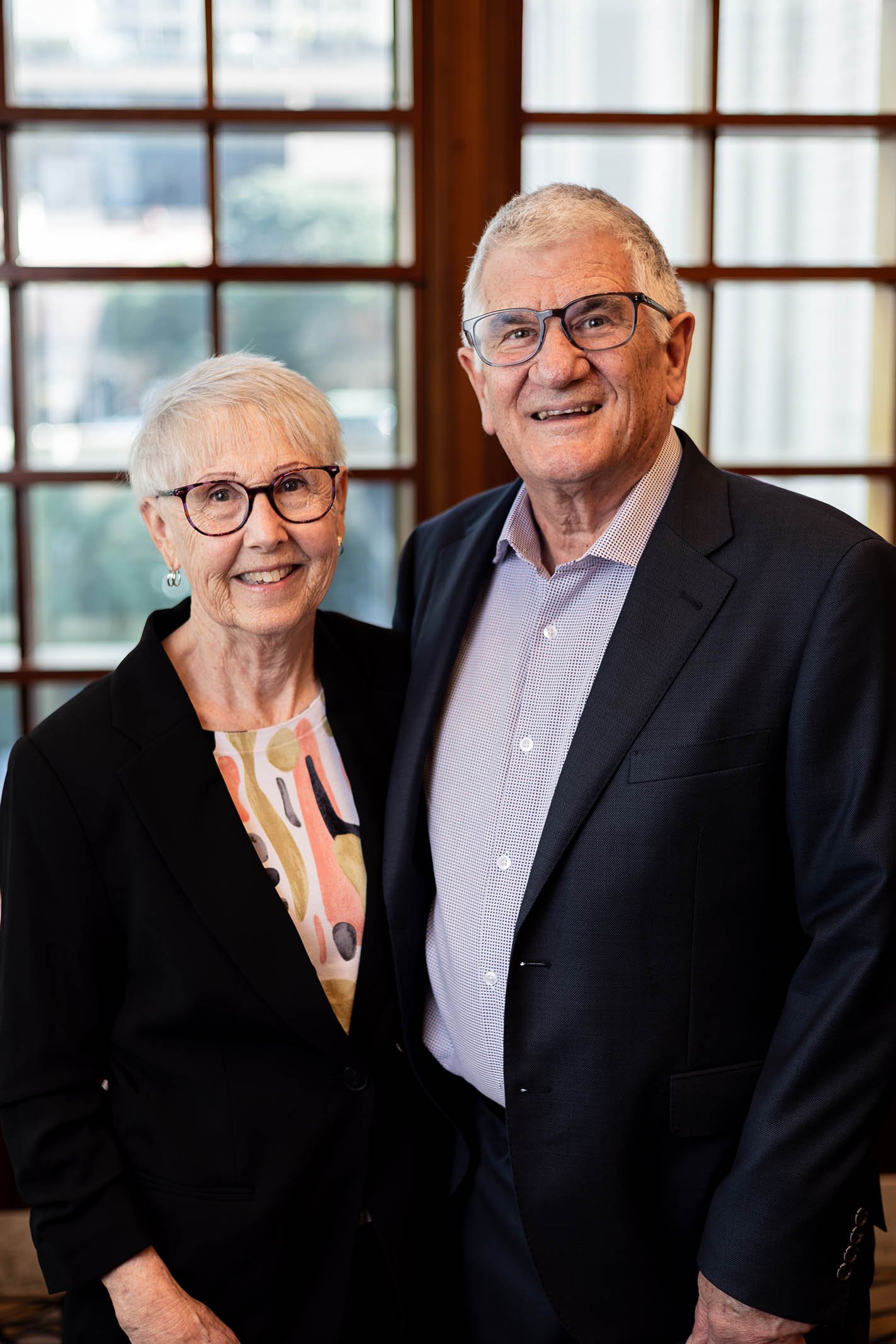 An elderly woman and an elderly man smiling and standing close together indoors near a large window.