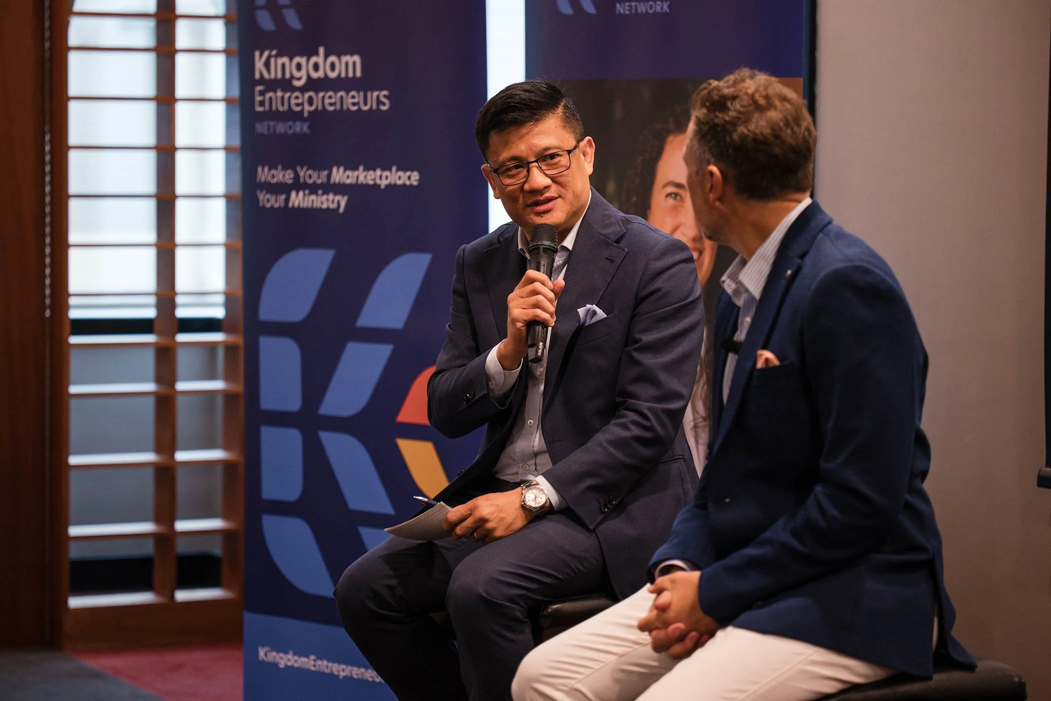 Two men in business attire sitting and talking at a professional event with a "Kingdom Entrepreneurs" banner in the background.