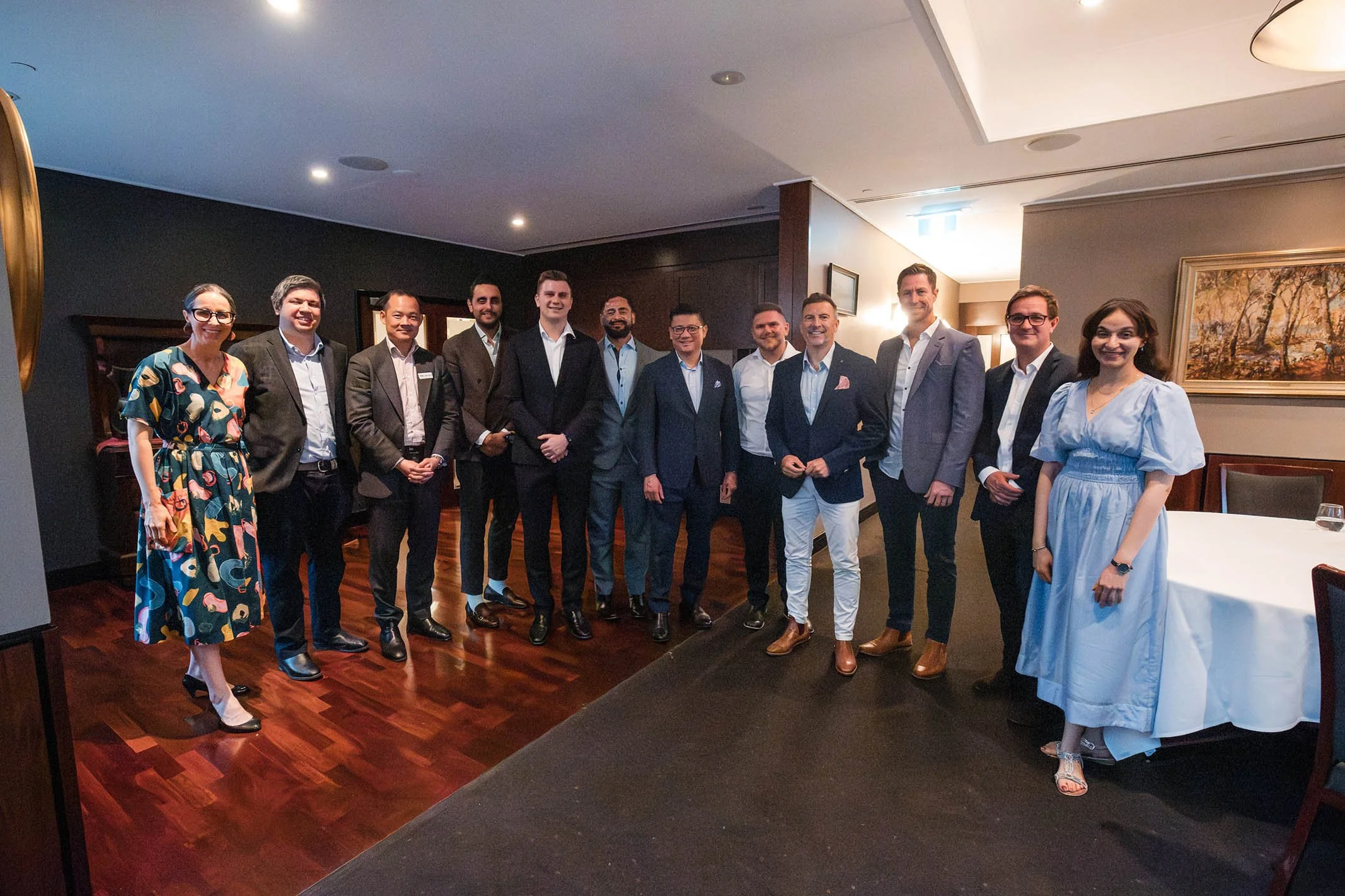 A group of fifteen professionally dressed individuals, including men and women, standing together in a well-lit, elegant event room with dark wood floors and artwork on the wall, posing for a photo at a formal gathering.