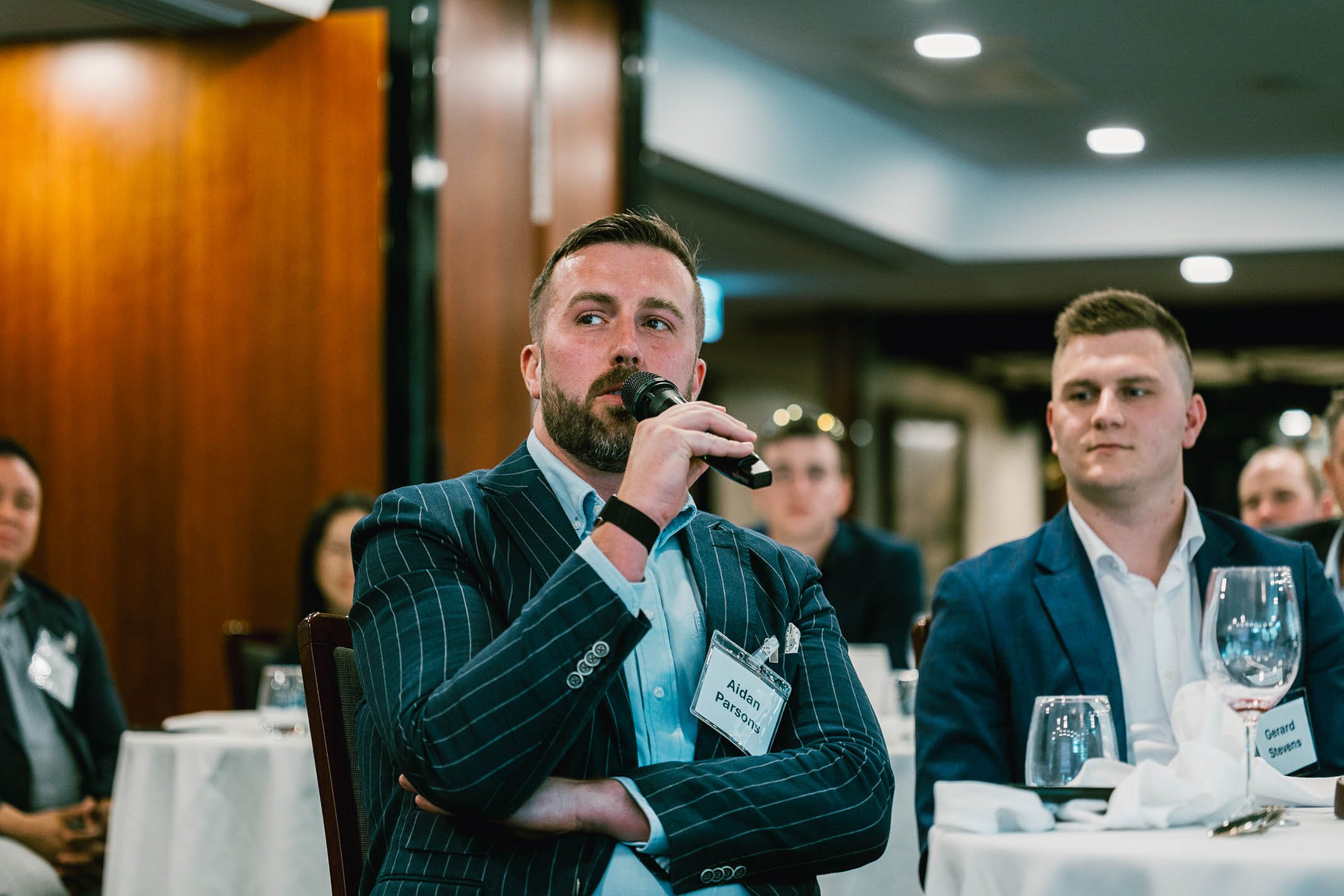 Man with a beard and short hair, wearing a blue striped suit, is speaking into a microphone at a conference or dinner event. He has a name tag that reads "Aidan Parsons." Several people are seated at round tables with white tablecloths, wearing busin