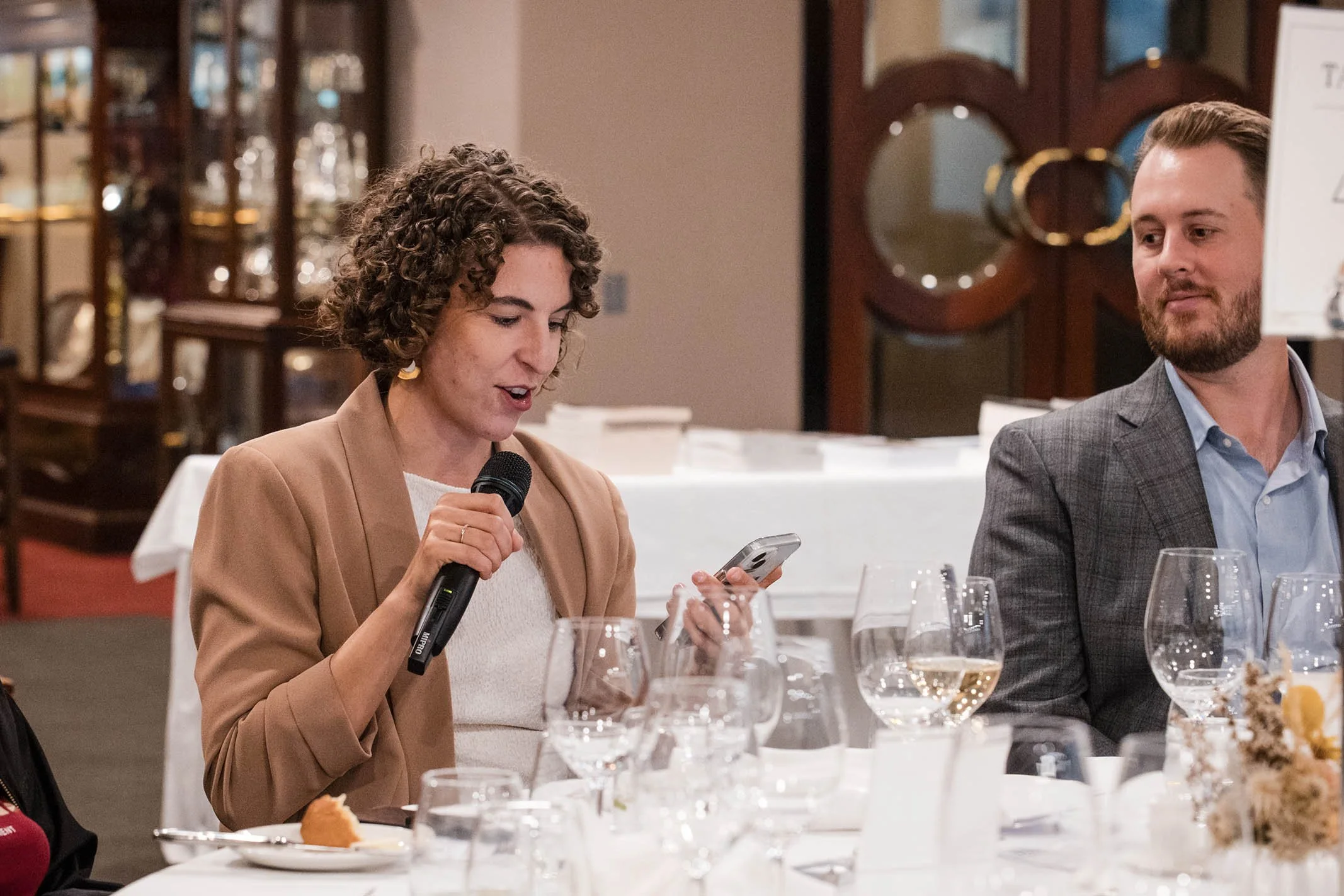 A woman with curly hair, wearing a tan blazer, holding a microphone and looking at her phone at a formal event or dinner. There are wine glasses and a table with a floral centerpiece in front of her, and a man in a gray suit sitting next to her.
