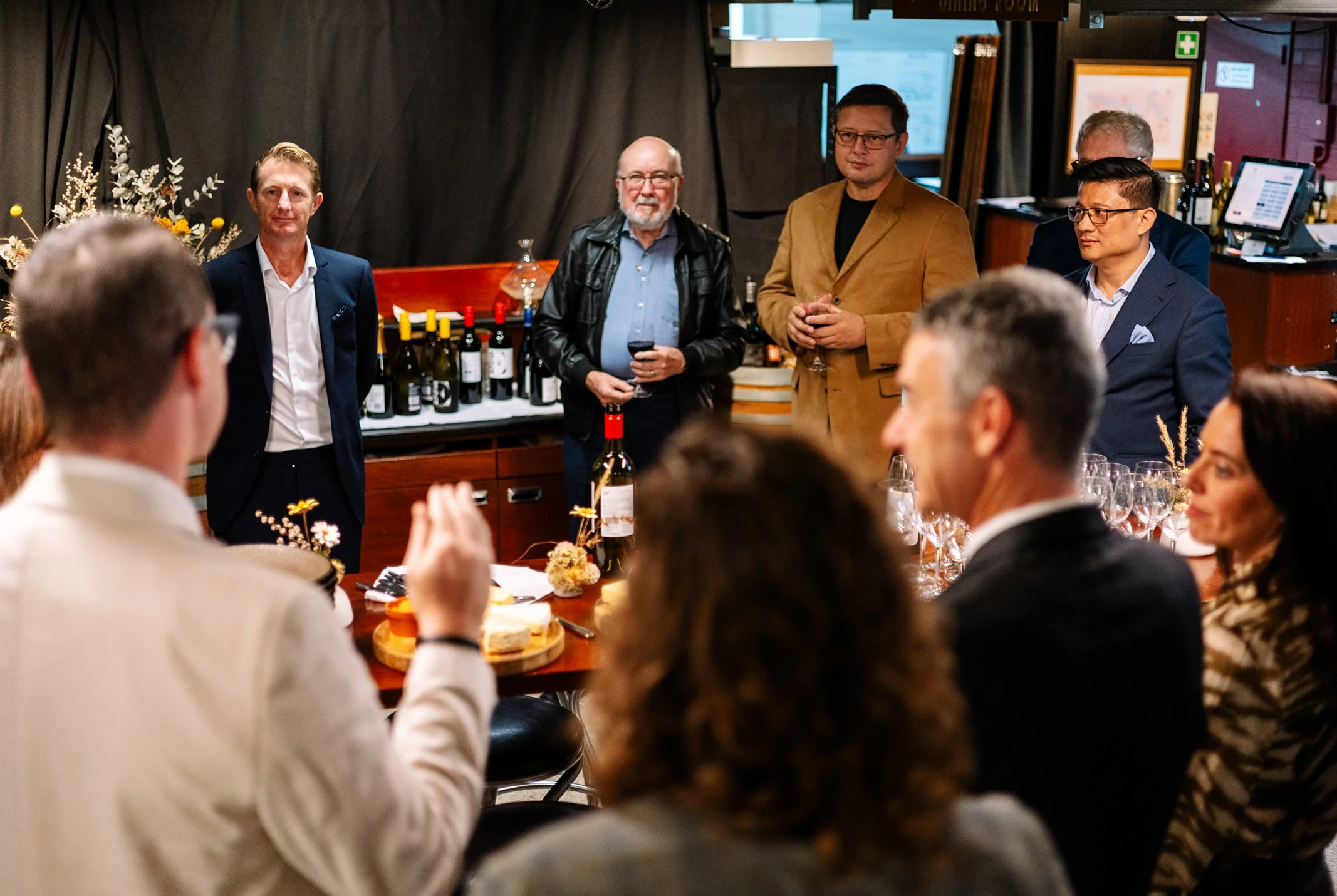 People gathering at a wine tasting event, with some standing and speaking, others seated around a table with wine glasses, wine bottles, and snacks, in a cozy indoor setting.