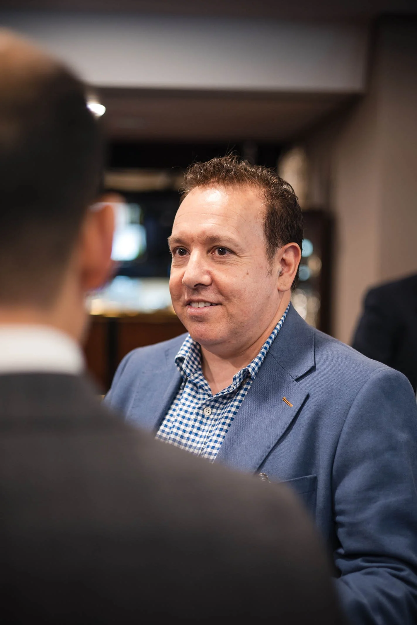 A man in a blue blazer and checkered shirt smiling and talking to someone in a professional setting.