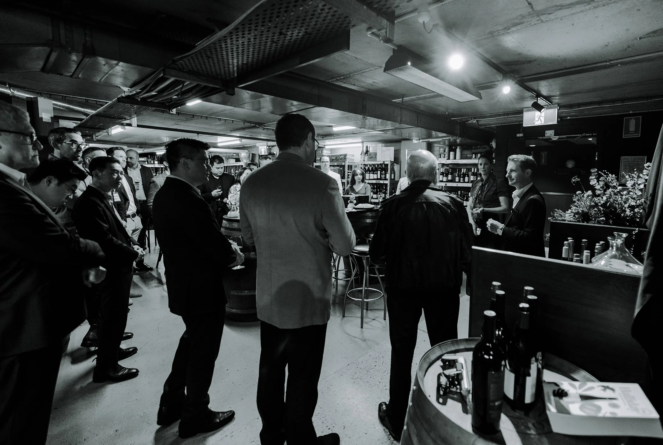 Group of people gathered in a bar or restaurant, some listening to a man speaking, with bottles of wine on shelves in the background, black and white photo.