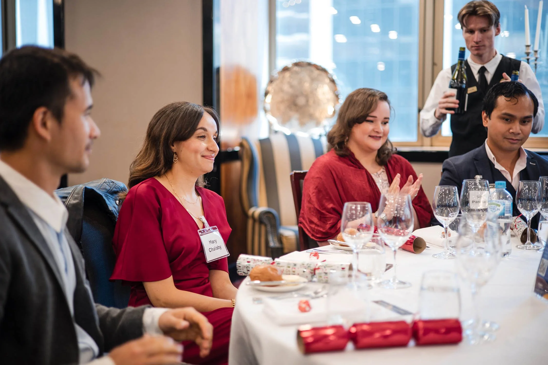 People sitting at a dinner table with onlookers clapping, a waiter serving drinks in the background.
