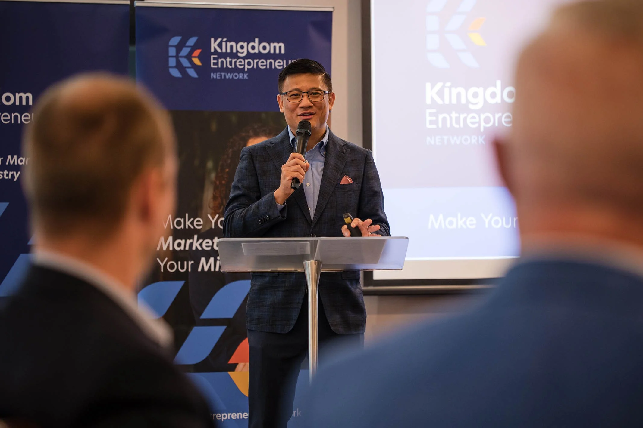 A man in a suit speaking at a podium during a conference with a Kingdom Entrepreneur Network banner in the background.