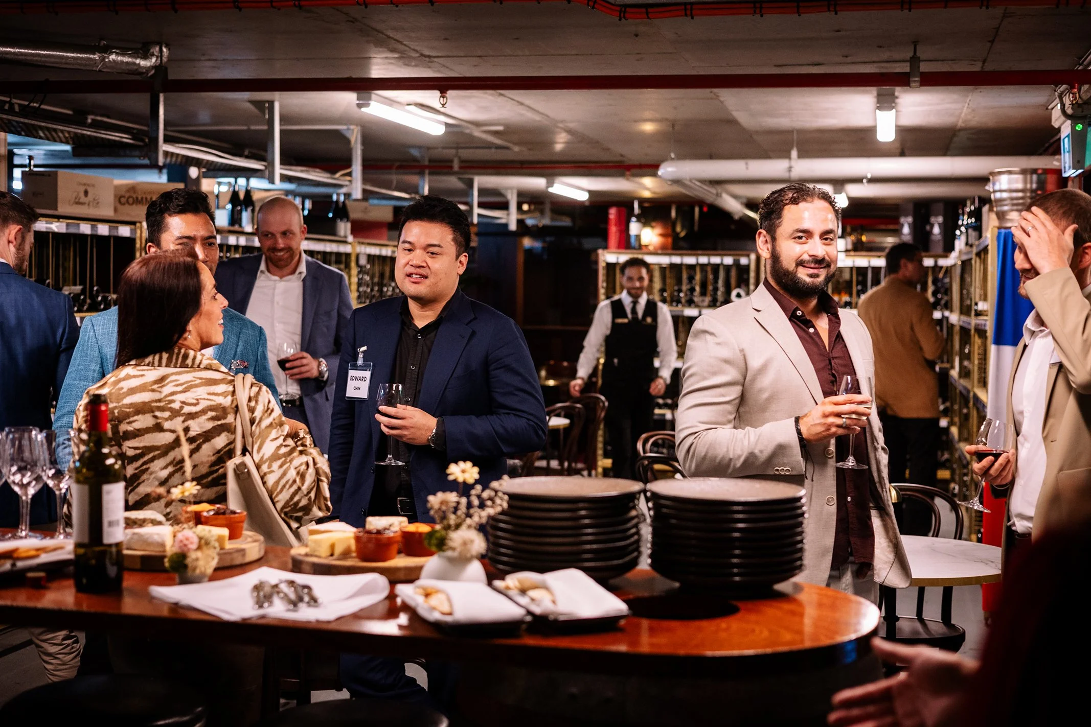 People socializing at a cocktail party in a modern venue with wine bottles, glasses, and catered food on a wooden table.