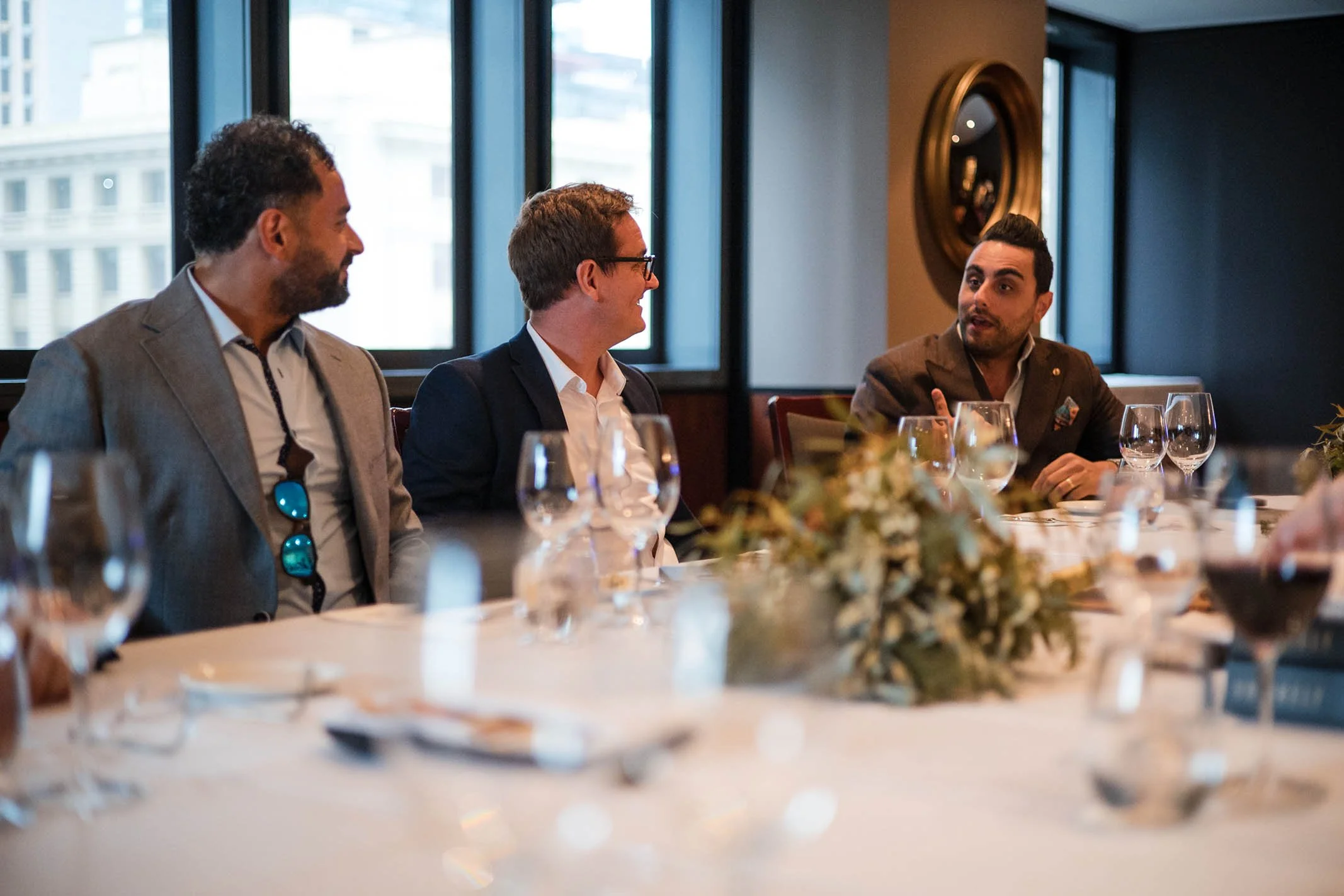 Three men are sitting at a dining table in a restaurant, engaged in conversation. The table is set with wine glasses, plates, and a floral centerpiece. The restaurant has large windows and modern decor.