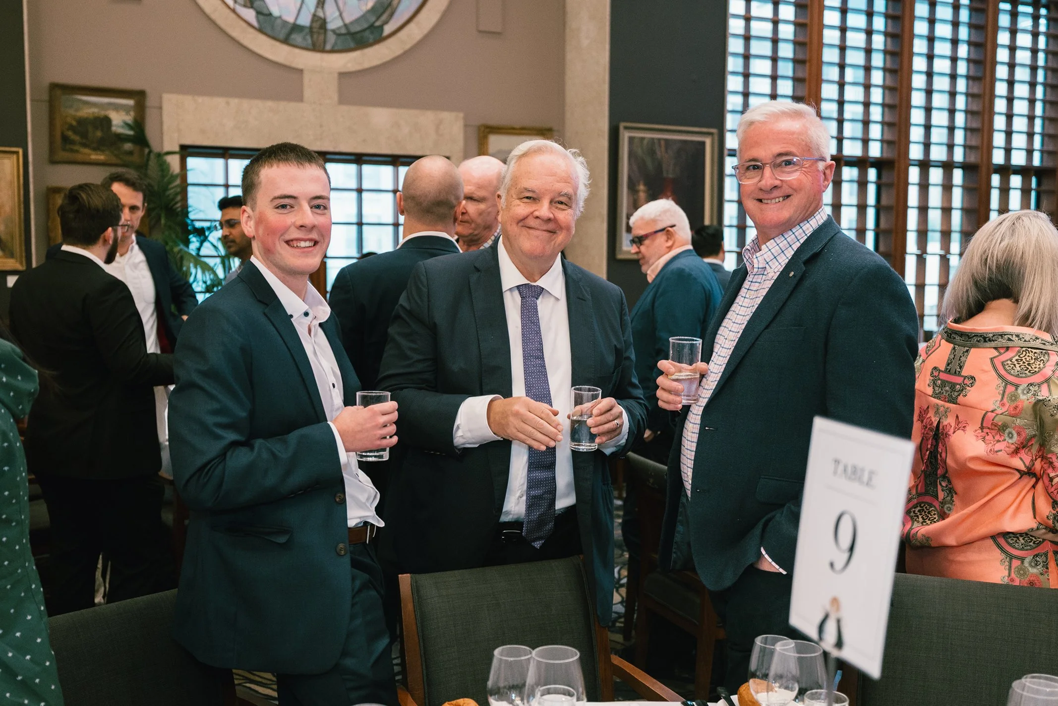 Three men in suits and ties at a formal gathering, holding glasses of water and smiling, with a crowded room and artwork in the background.