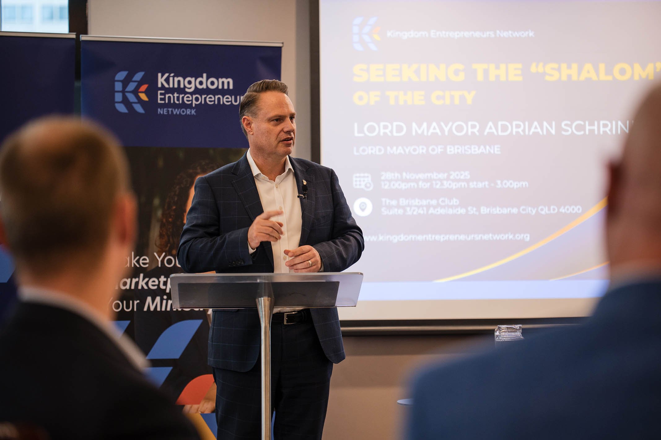 Lord Mayor Adrian Schrin giving a presentation at a conference, with a banner and a screen displaying event details.