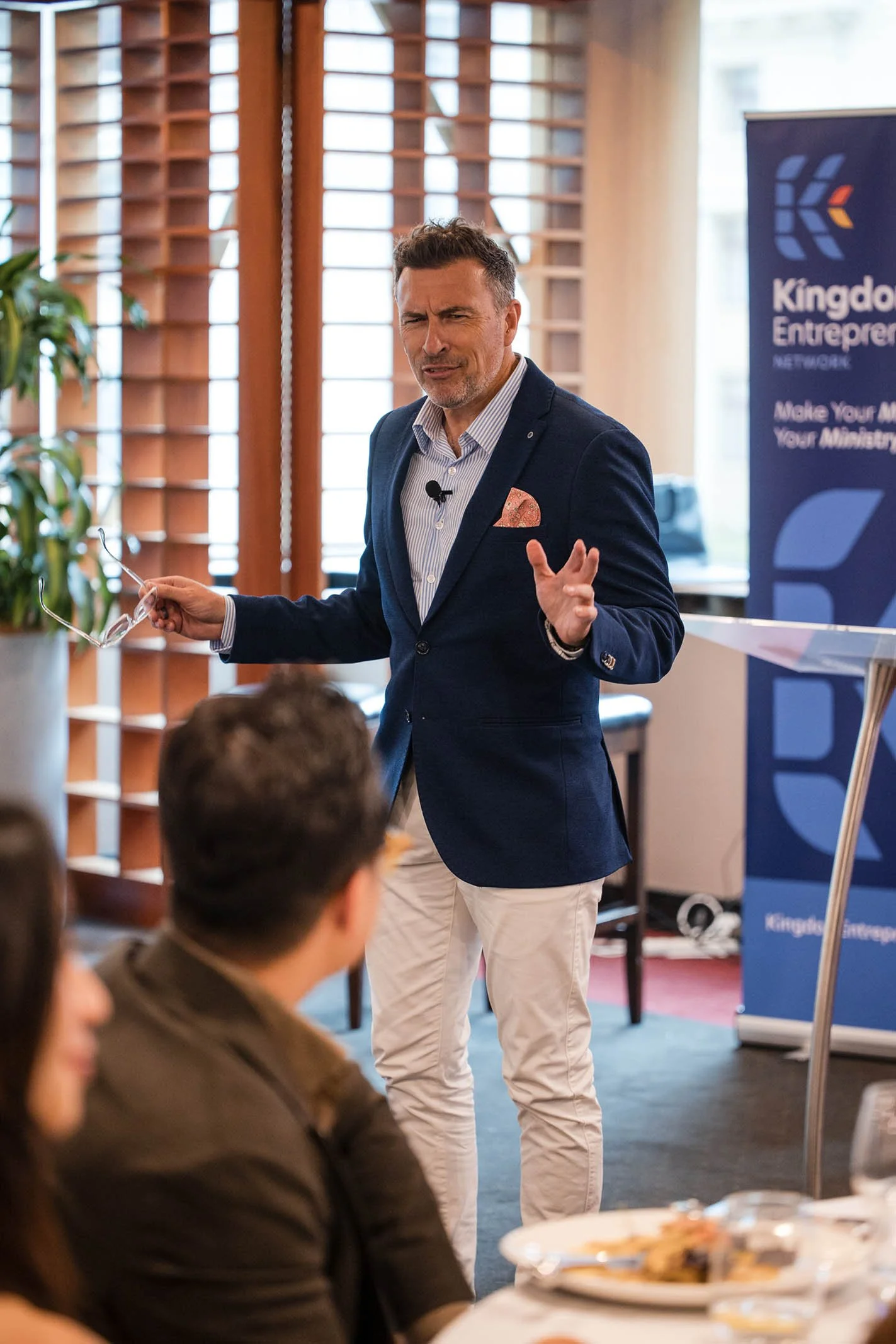 A man in a blue blazer and white pants giving a presentation in a conference room. He holds glasses in one hand and gestures with the other. There is a banner in the background with the text 'Kingdom Entrepreneurship Network'. Several people are seat