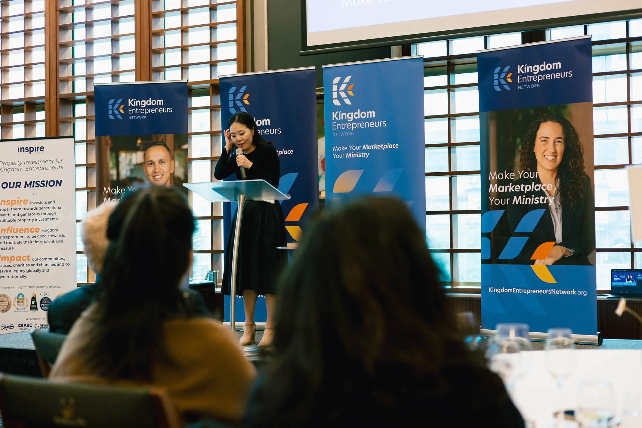 A woman giving a presentation at a conference with banners behind her reading 'Kingdom Entrepreneurs Network' and 'Make Your Marketplace Your Ministry' in a well-lit room with large windows, audience members seated in front.