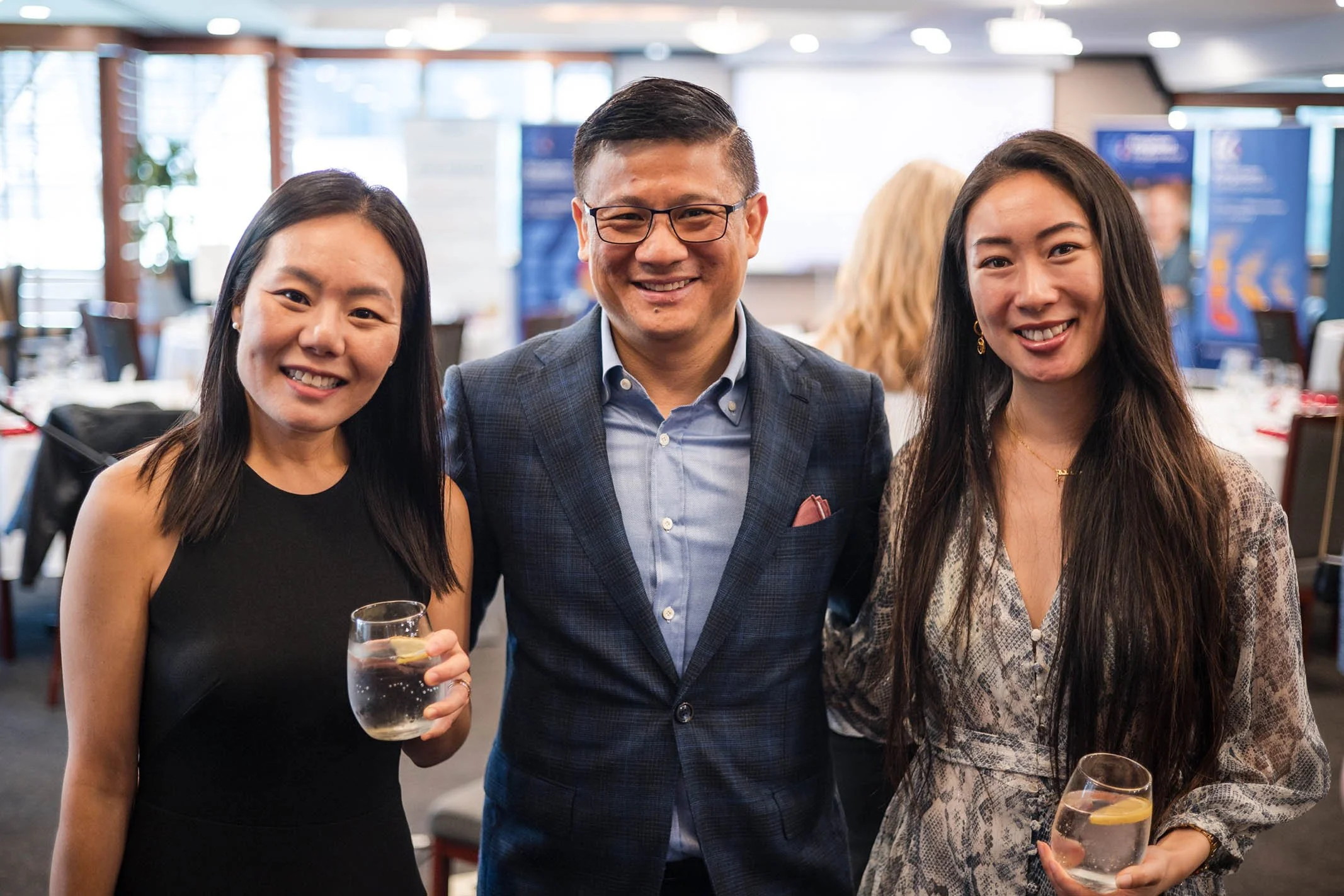 Three people smiling at a social event, two women holding glasses of water with lemon, a man in the middle wearing glasses and a blazer.