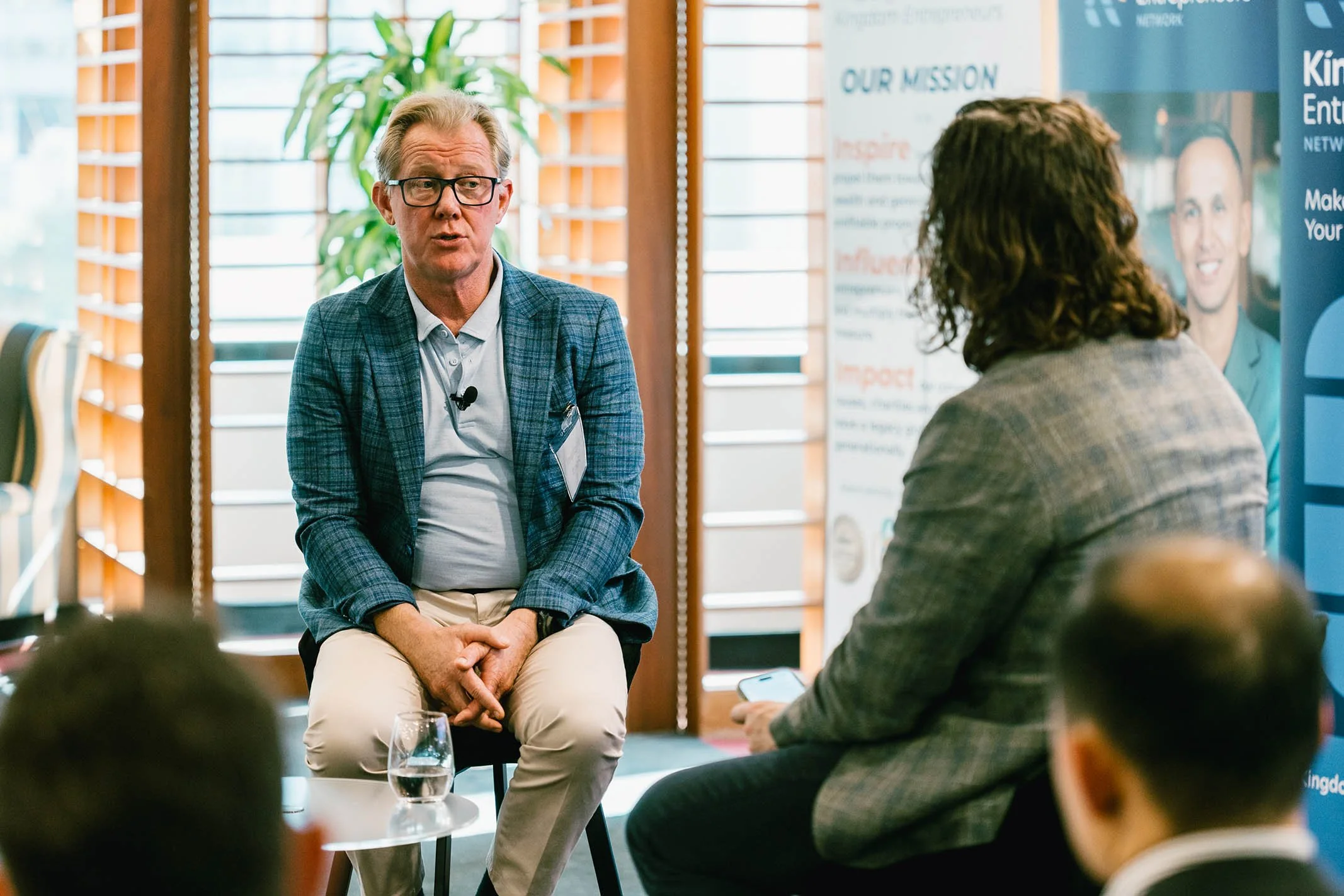 A man in glasses and a plaid blazer is speaking in a conversation, sitting on a chair with a glass of water in front of him. A woman with curly hair, wearing a gray blazer, is listening and holding a phone. There are blurred people and informational 