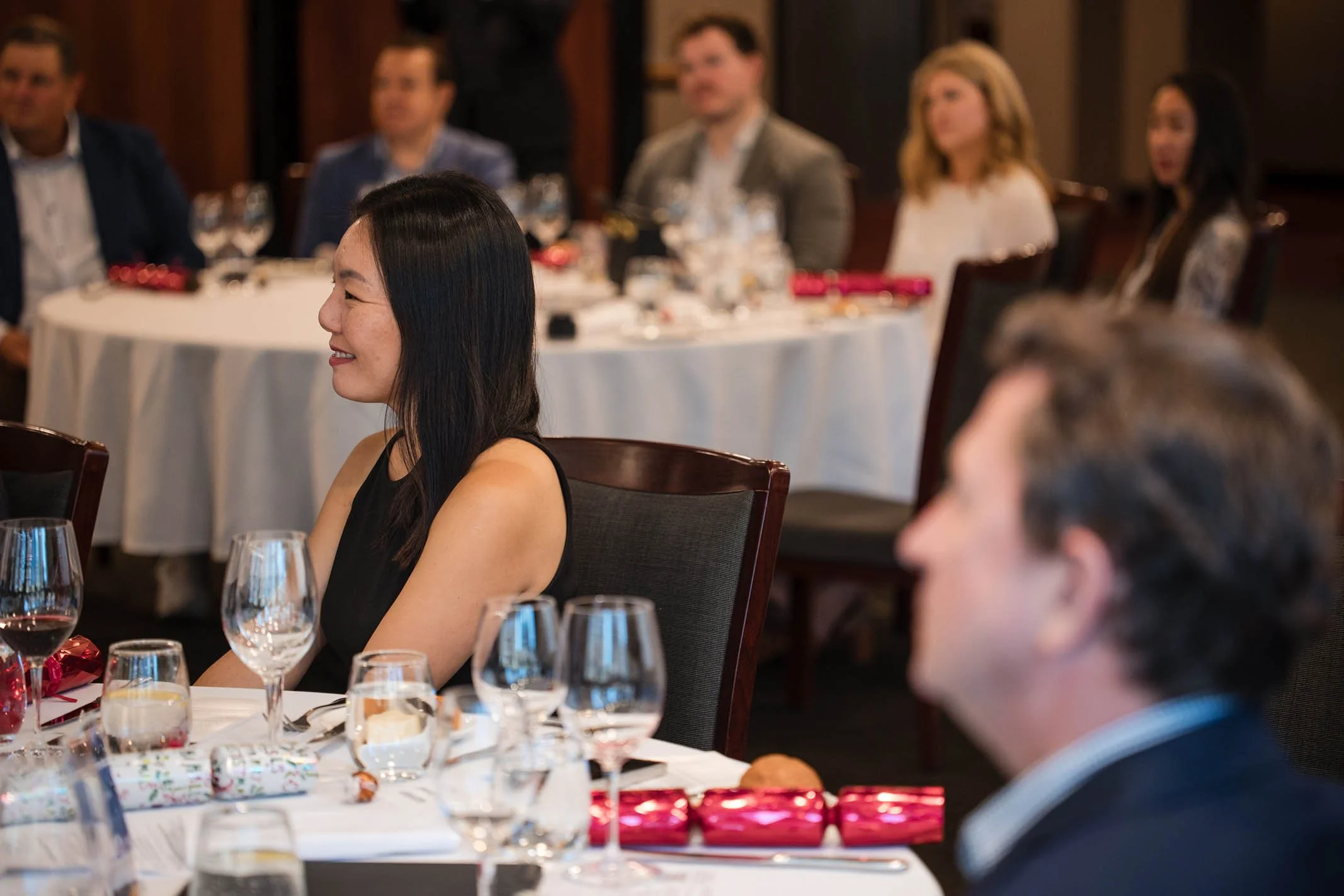 People sitting at round tables with white tablecloths, wine glasses, and red wrapped gifts, attending a formal event or dinner party in a banquet hall.