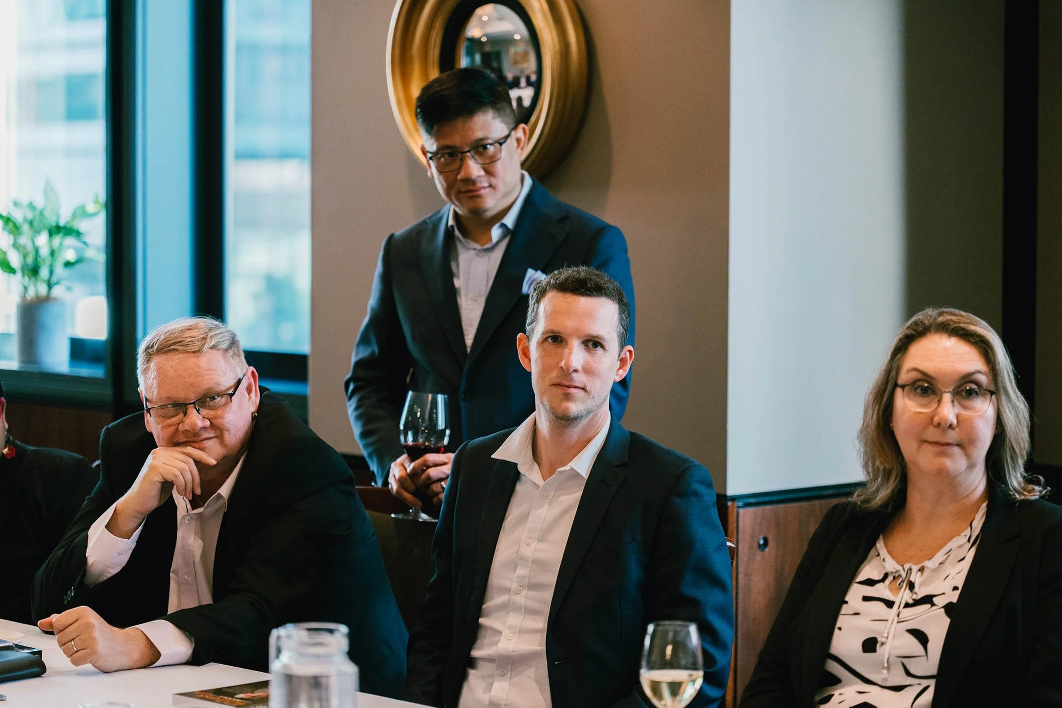 Four people attending a formal meeting or event, seated at a table, with one standing behind them. They are dressed in business attire and appear to be listening or waiting. The background shows a window with natural light and a decorative mirror on 