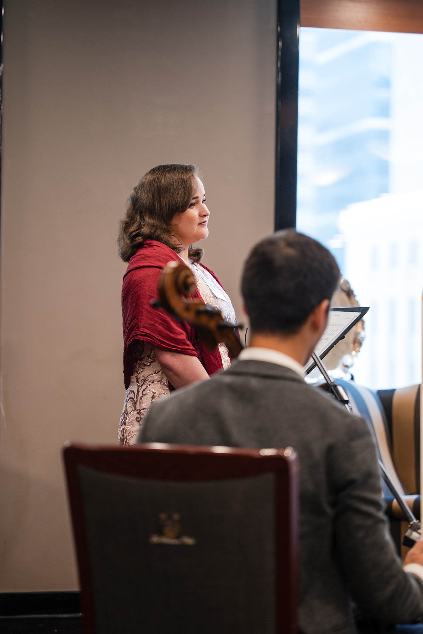 A woman with brown hair, wearing a red shawl and patterned dress, stands playing a violin during a performance. A man in a gray suit is sitting in front of her with his back to the camera, reading sheet music, with another musician partially visible 