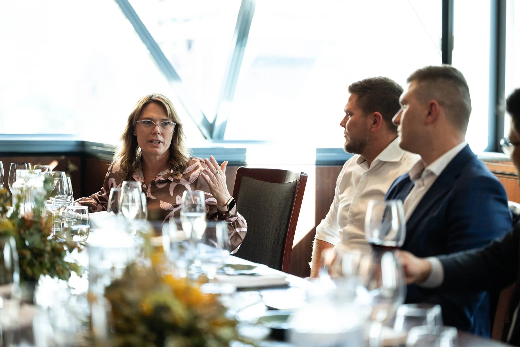A woman is speaking and gesturing with her hand during a meeting with three men sitting at a table with glasses and plates in a bright room with large windows.