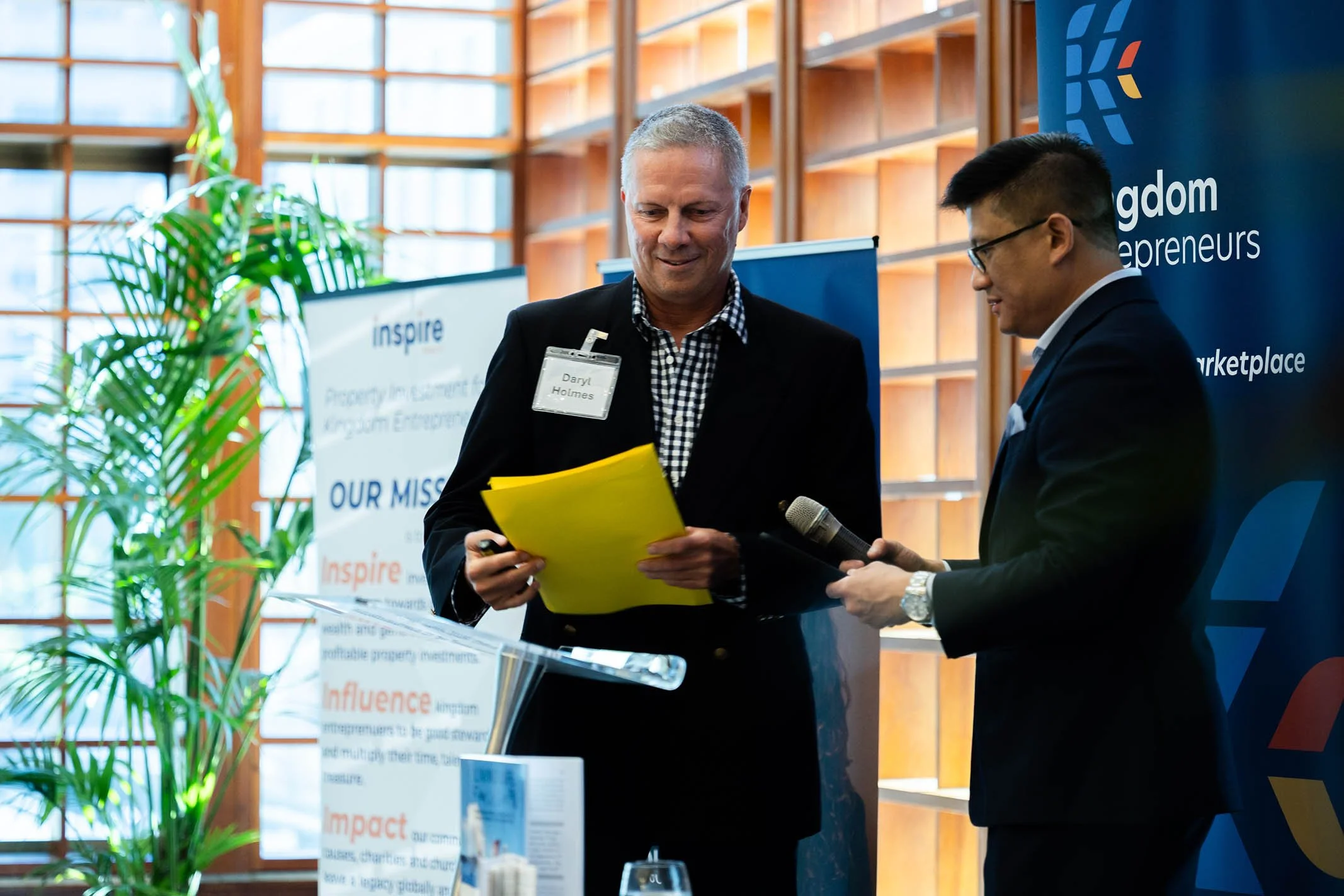 Two men in suits at a professional event, one is holding a yellow folder and the other is holding a microphone, with banners and a plant in the background.
