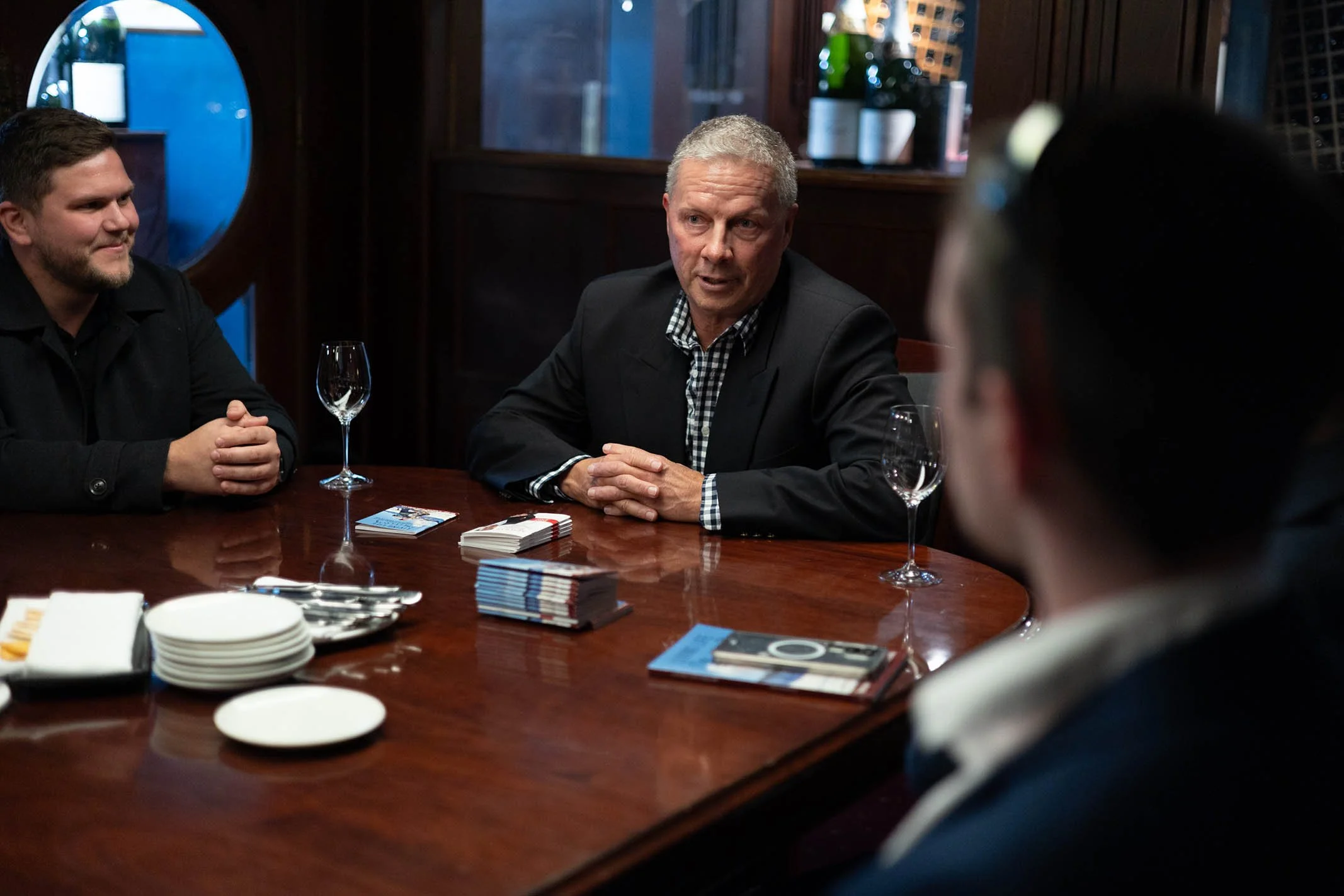 Three men sitting at a wooden table engaged in conversation, with cards, magazines, plates, and wine glasses on the table, in a dimly lit room with dark wood paneling.