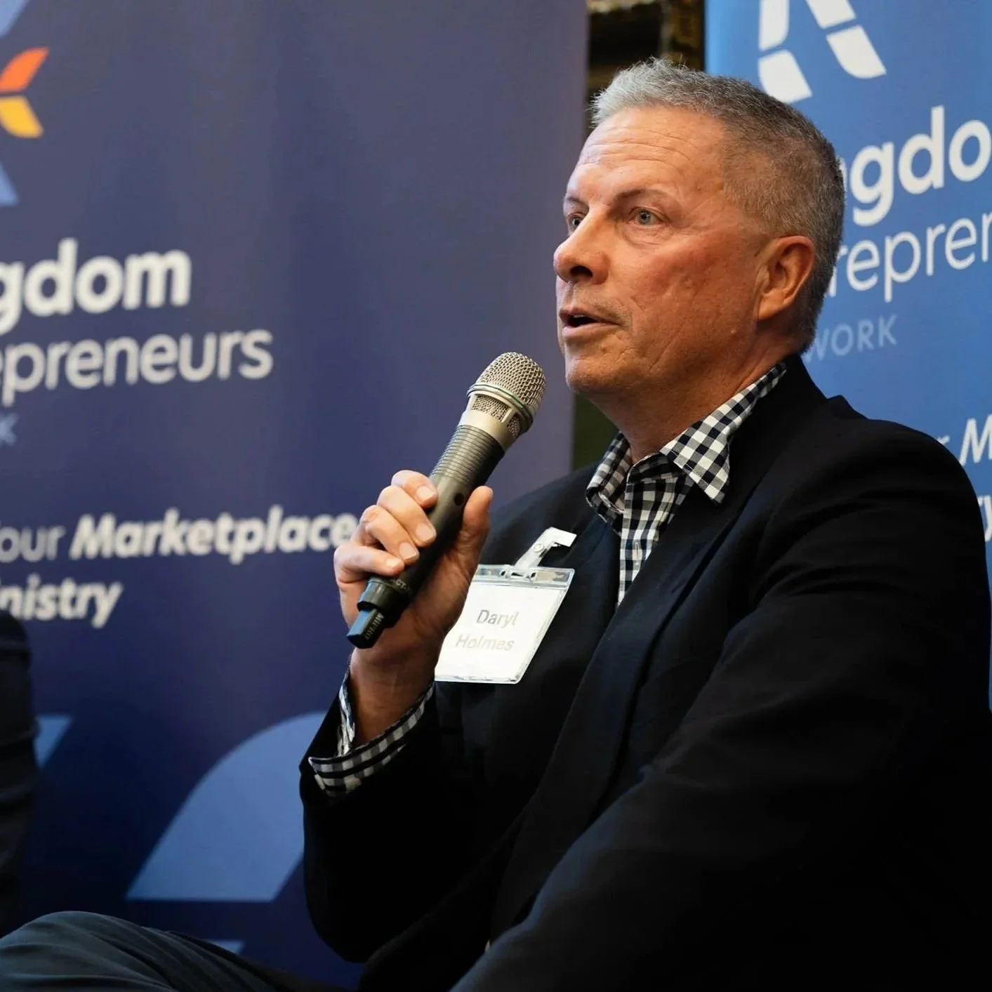 A middle-aged man with short gray hair, wearing a black blazer and a checkered shirt, speaking into a microphone at a professional event, with a blue backdrop that has white text and logos.