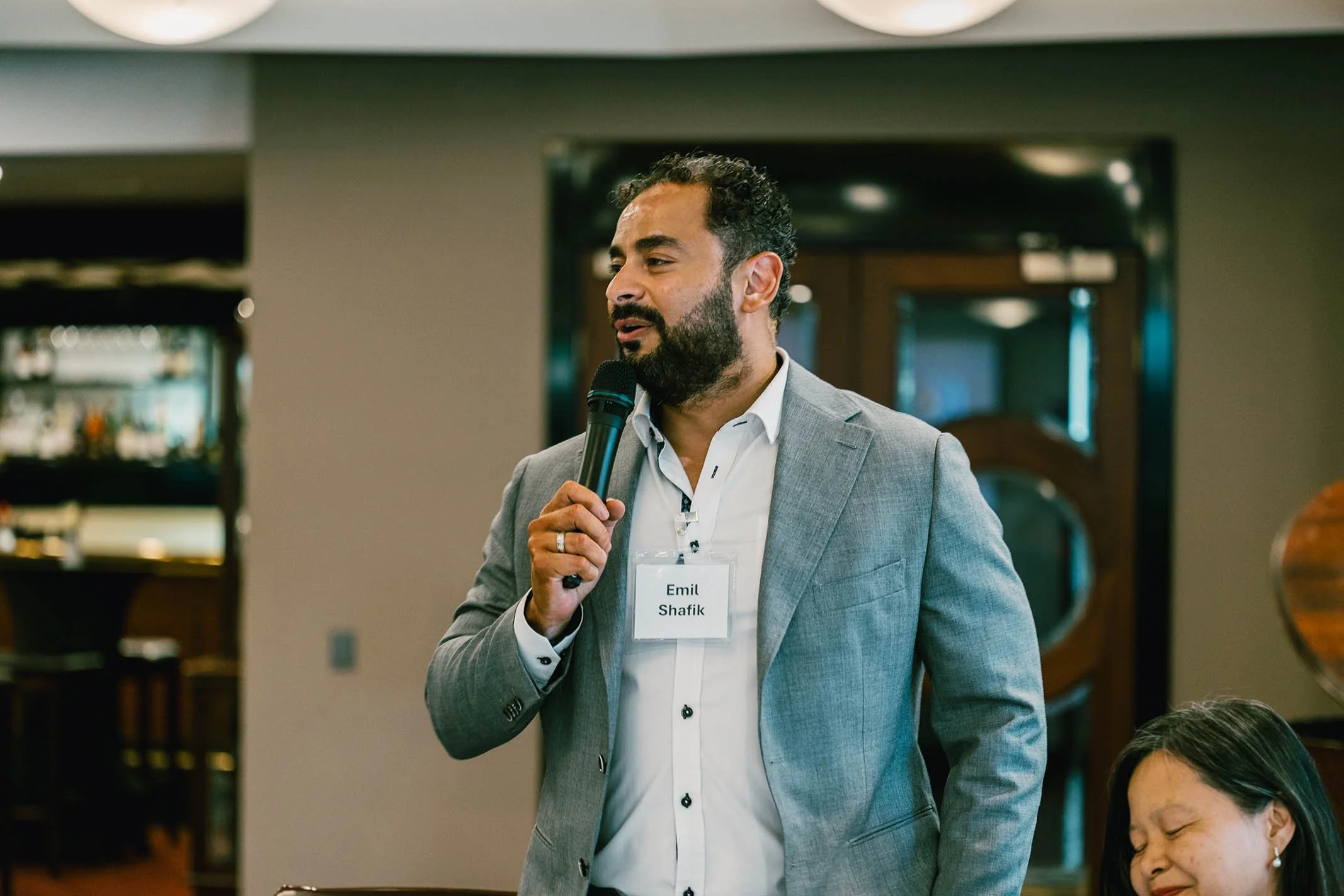 A man with a beard, wearing a gray suit and white shirt, holds a microphone and speaks at an event. He has a name tag that reads 'Emil Shafik'. A woman with long dark hair is sitting nearby.