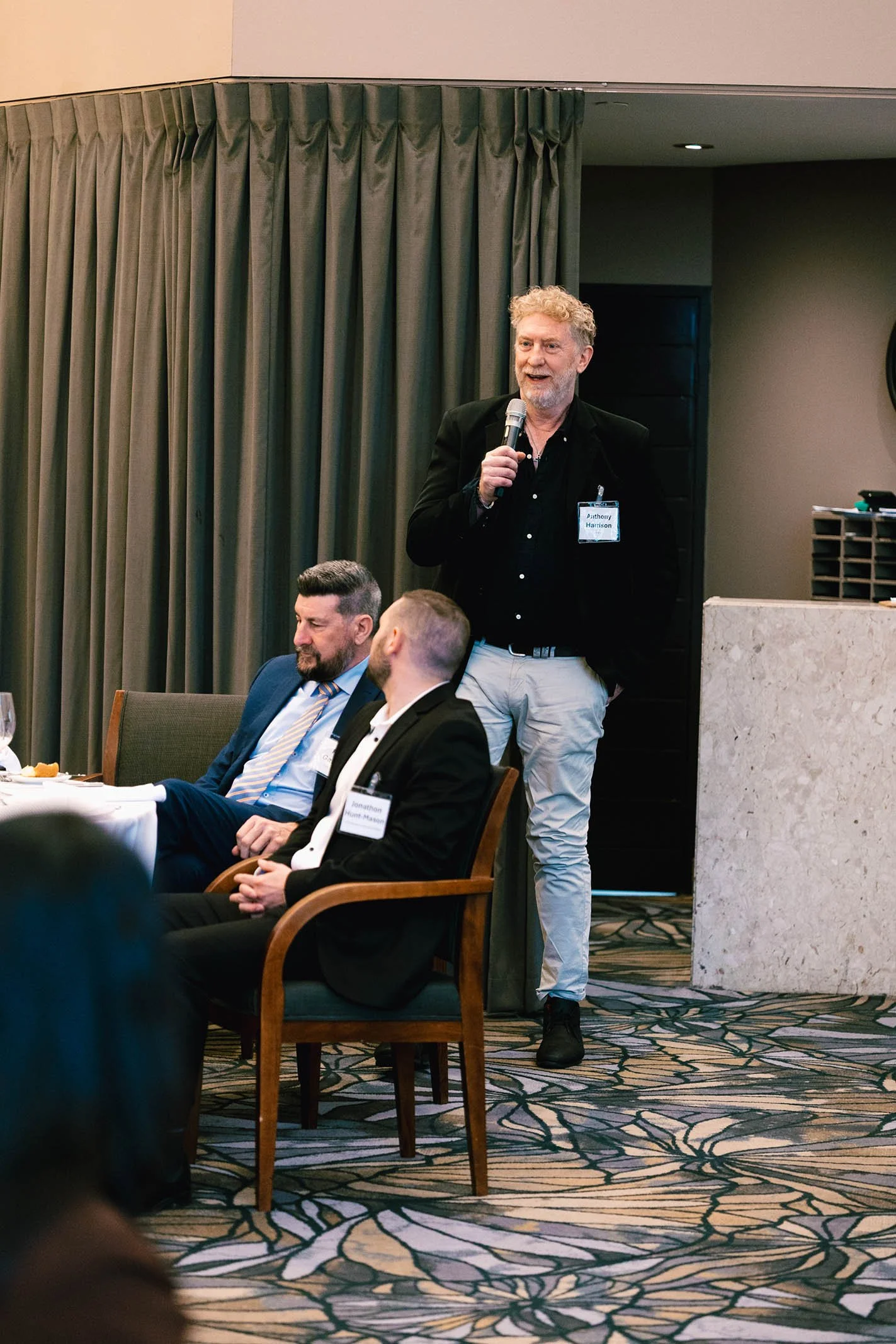 Man speaking into a microphone during a conference or meeting, seated at a table with two other men, in a room with beige walls and green curtains.