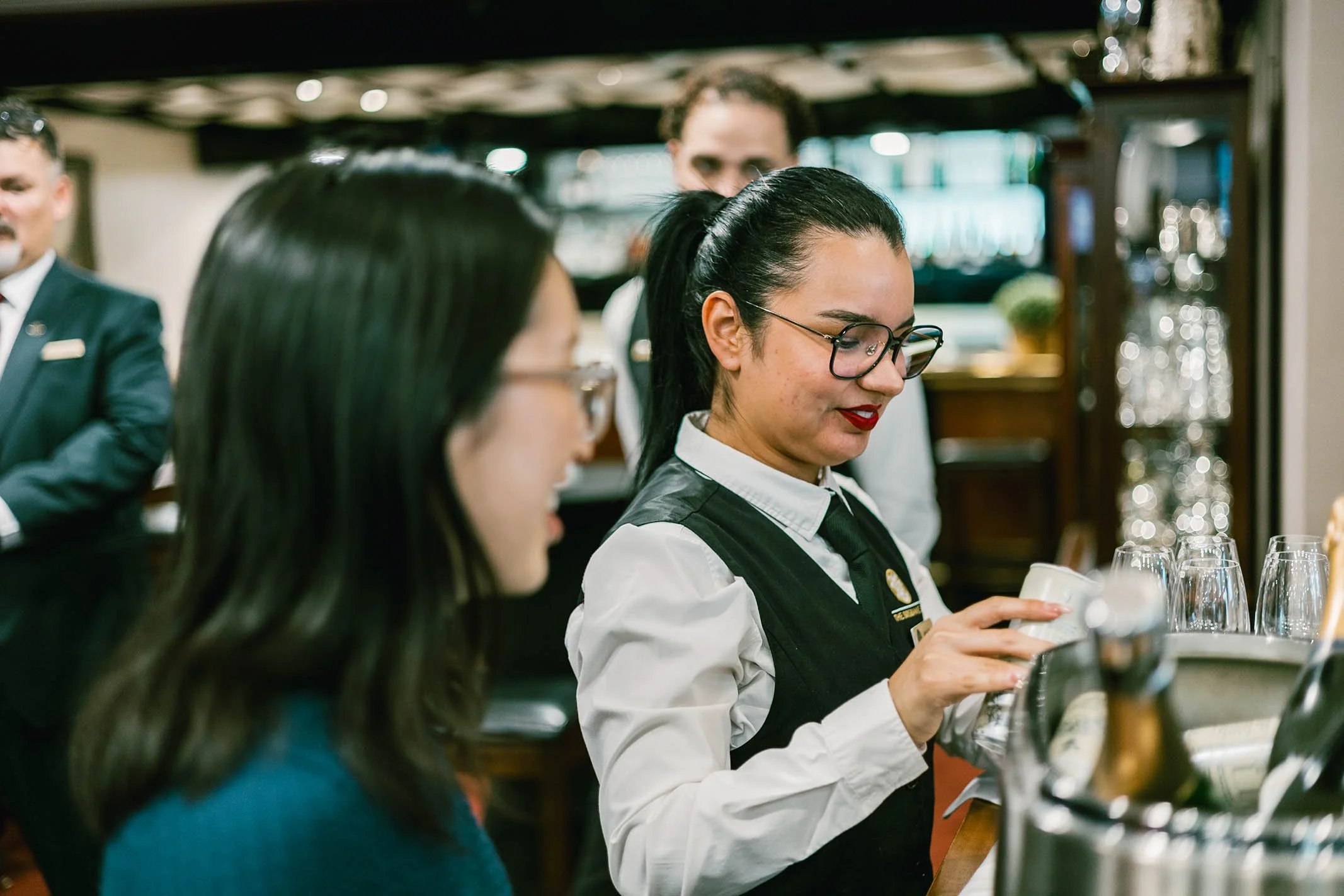 A waitress with glasses and dark hair tied back, wearing a white shirt and black vest, serving drinks to a woman with glasses and long dark hair, in a restaurant or bar setting.