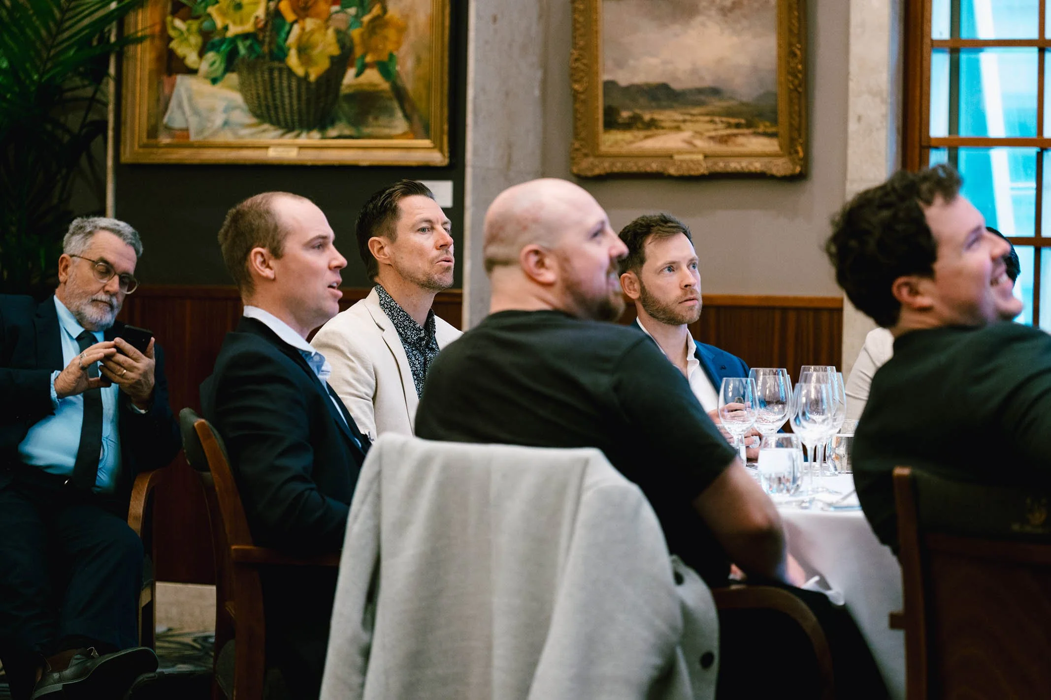 A group of men seated at a formal event in a room with framed paintings on the wall, some focusing on a presentation or speaker, one using a phone, and several with empty wine glasses in front of them.