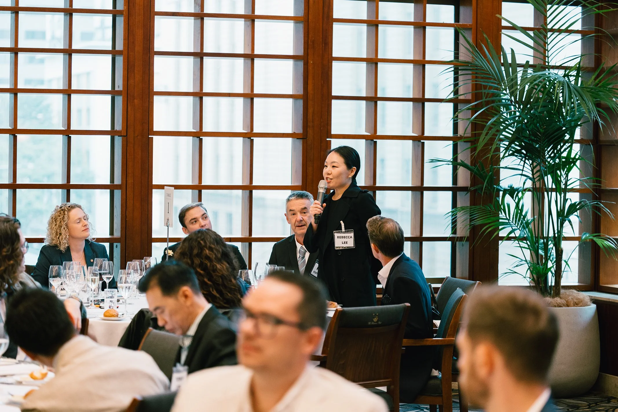 A woman with a microphone speaking at a formal event with seated attendees listening. The setting features large wooden grid windows and tall potted plants.