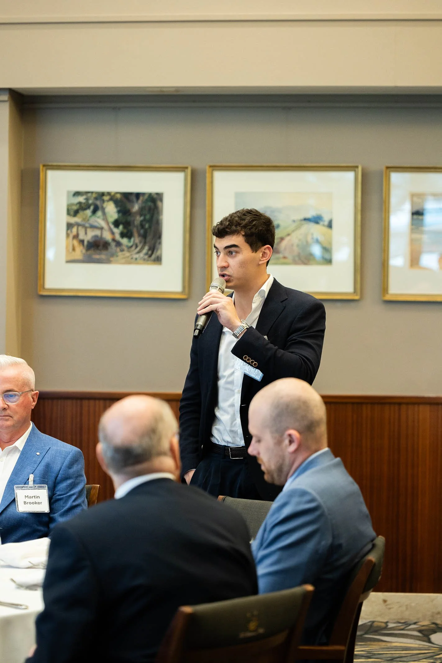 A young man in a business suit standing and speaking into a microphone during a formal meeting or conference. Several seated middle-aged men are visible, listening. The background features framed landscape paintings on a gray wall.