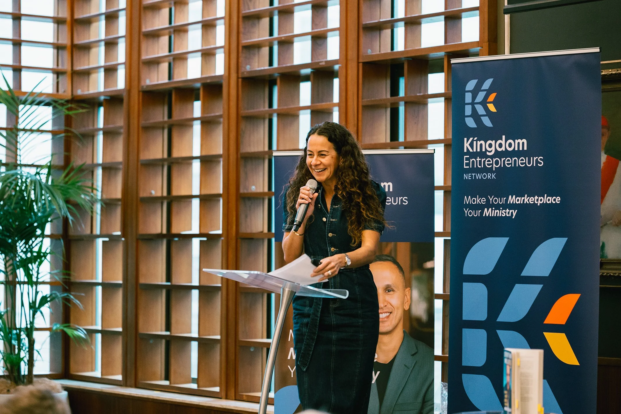 A woman with long, curly hair speaking into a microphone at a lectern during a conference or event, with banners for 'Kingdom Entrepreneurs Network' behind her, in a room with wooden paneling and large windows.
