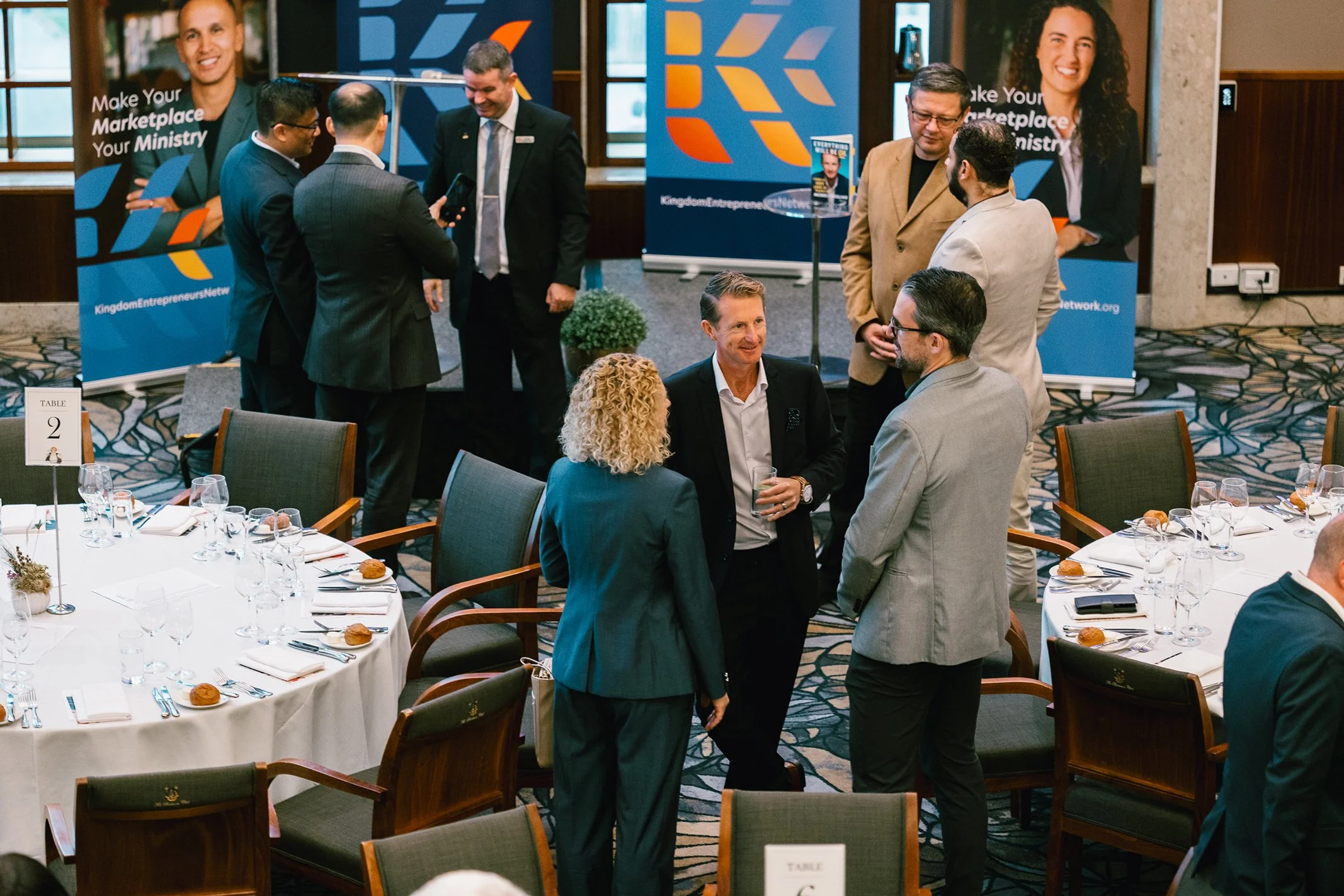Group of professionals socializing and networking at a formal event or conference, with round tables set with glassware and place settings, in a decorated banquet hall.