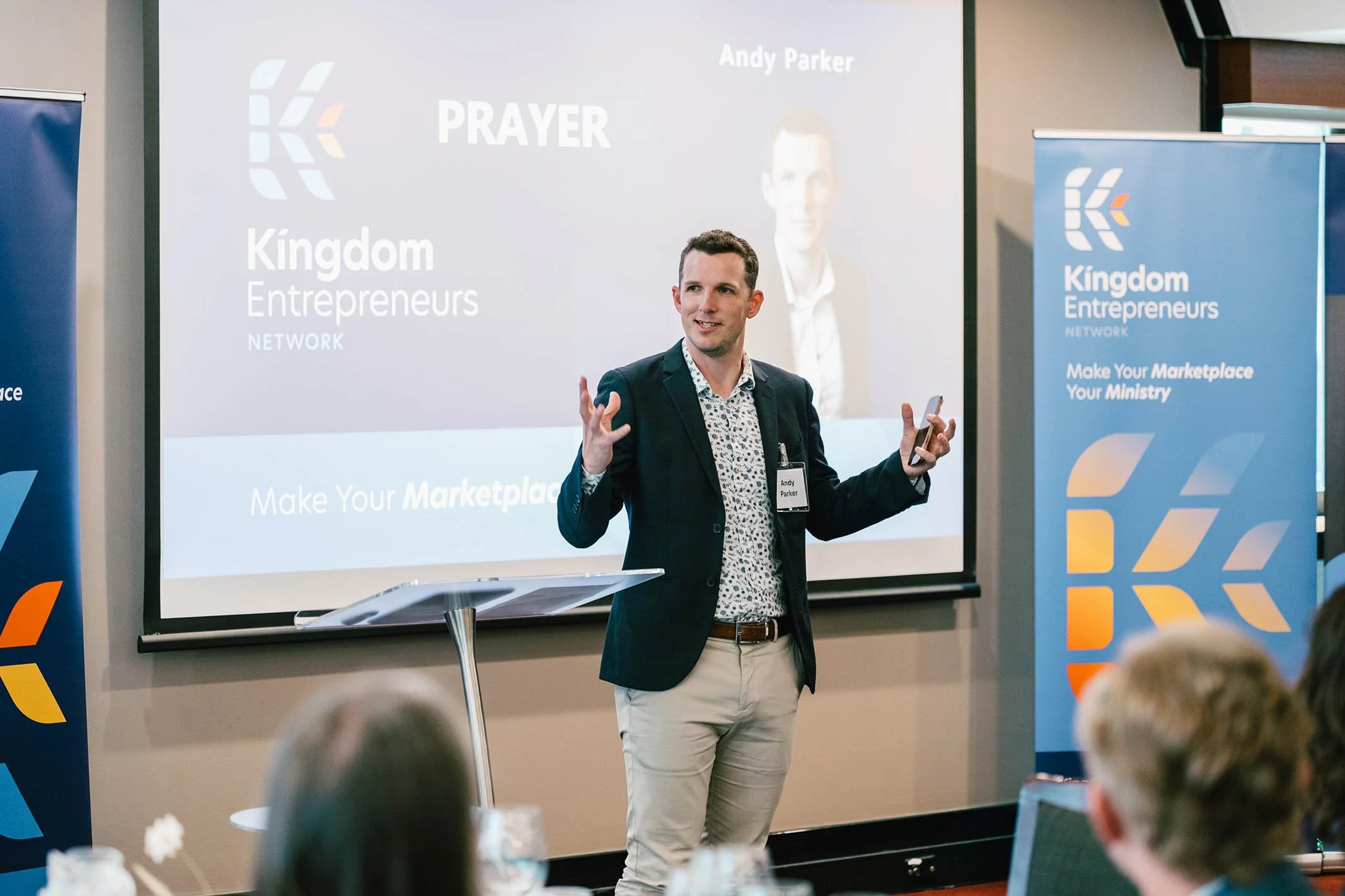 A man in a dark blazer and patterned shirt giving a presentation at a conference, with a screen and banners displaying 'Kingdom Entrepreneurs' behind him.