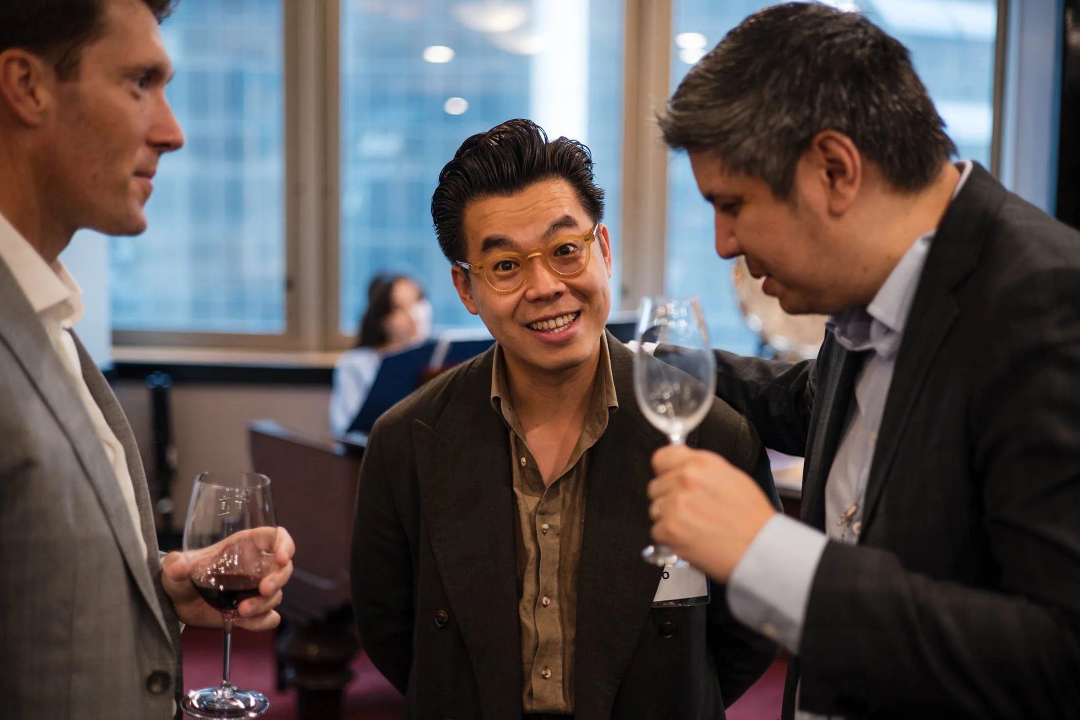 Three men in business attire talking and smiling at a social gathering in a modern office or conference room, with two of them holding glasses of wine.