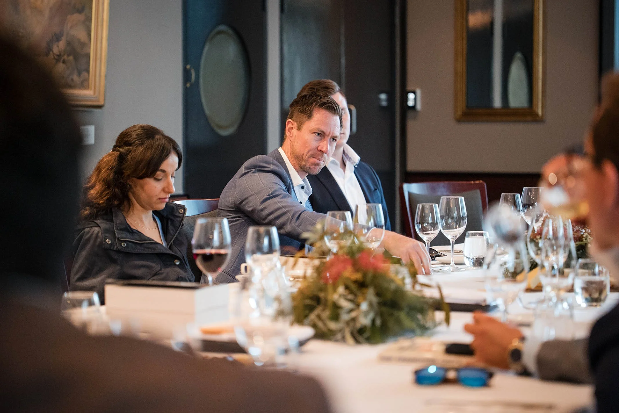 A group of people seated around a conference table during a meeting, with glasses of wine and water, and a floral centerpiece in the middle, in a professional setting.