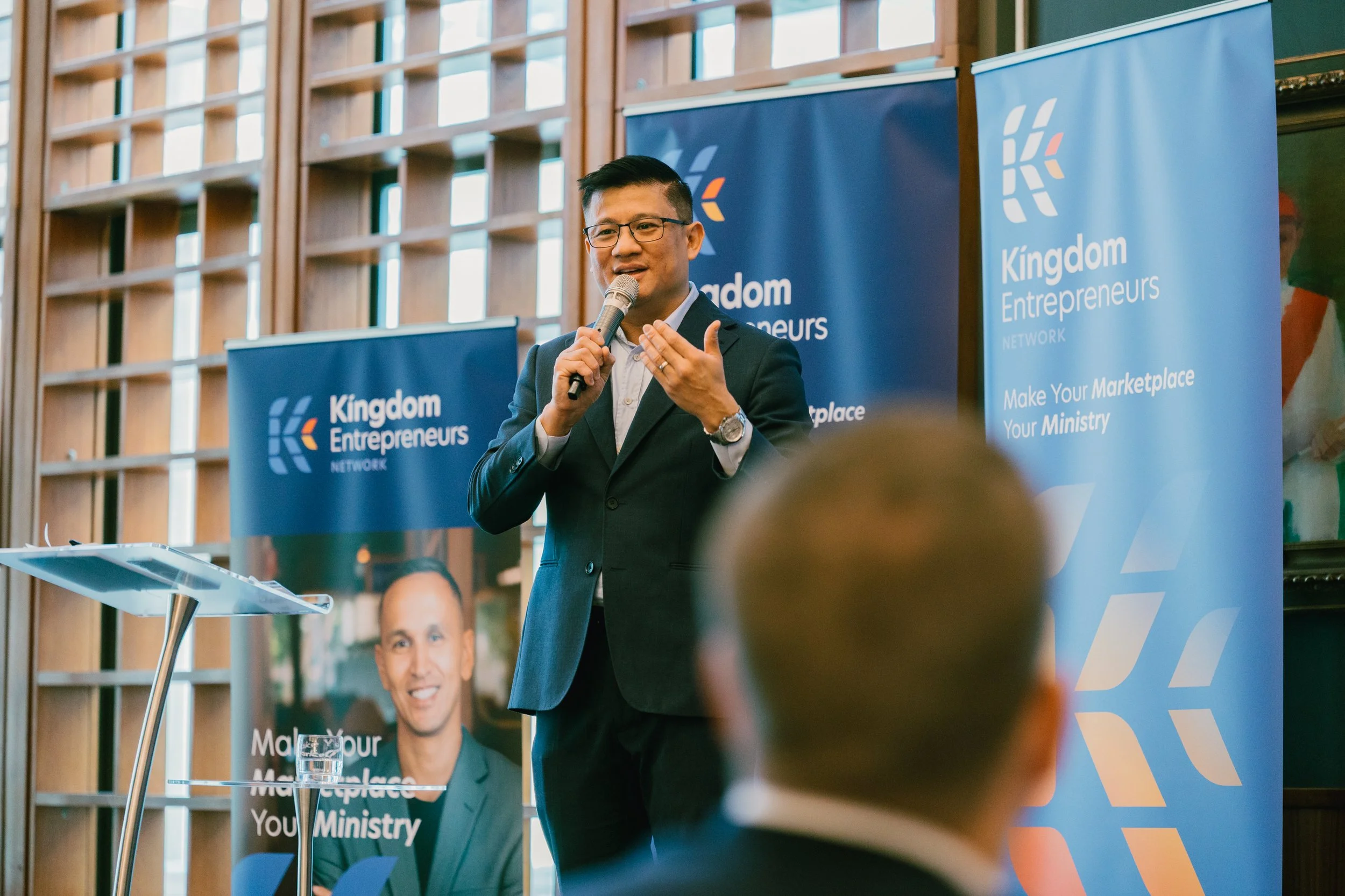 A man in a dark suit speaking into a microphone during a presentation at the Kingdom Entrepreneurs Network event, with banners in the background that read "Kingdom Entrepreneurs NETWORK" and "Make Your Marketplace Your Ministry."