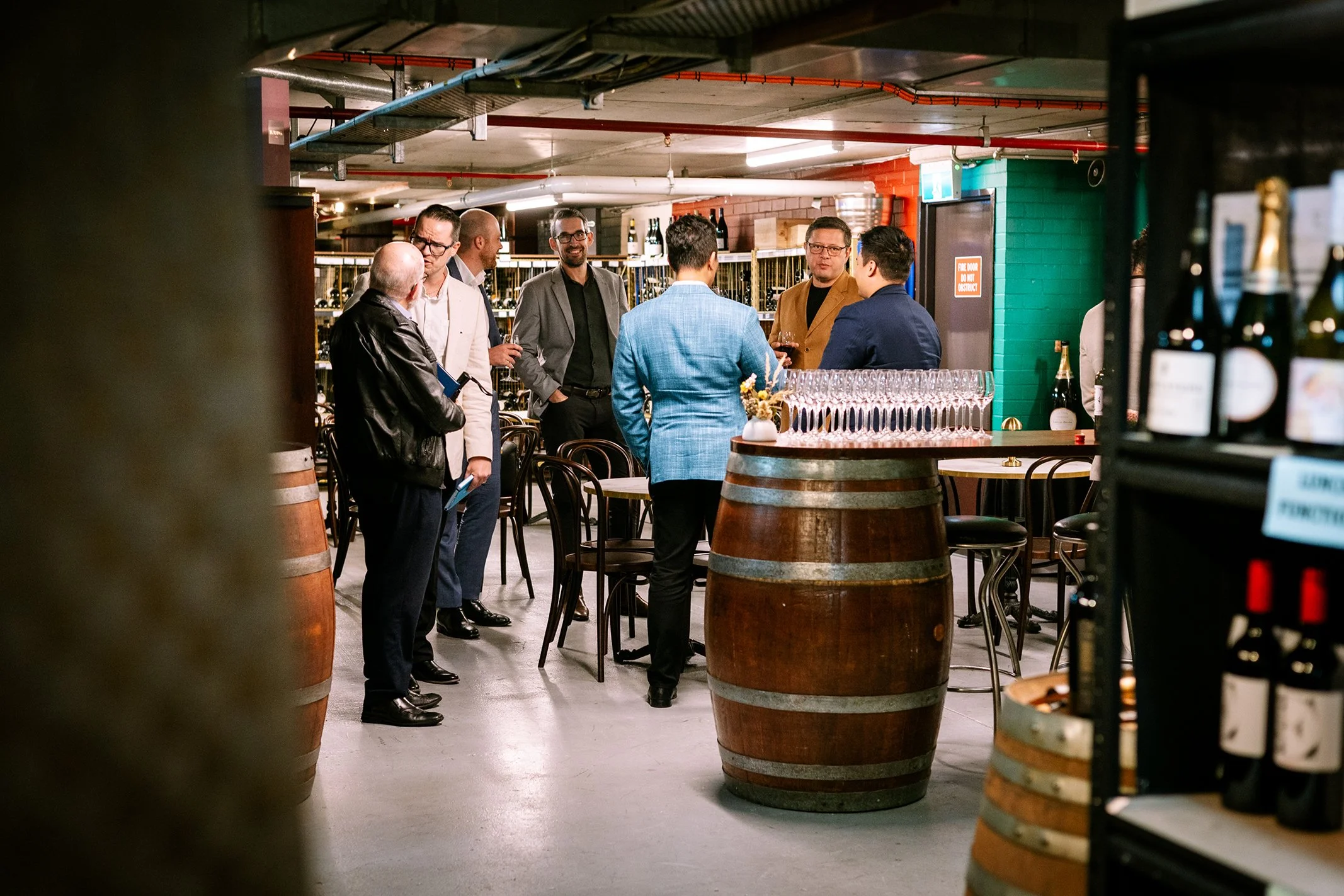 Group of casually dressed men socializing around a barrel table in a wine cellar or tasting room, with bottles of wine and glasses around.