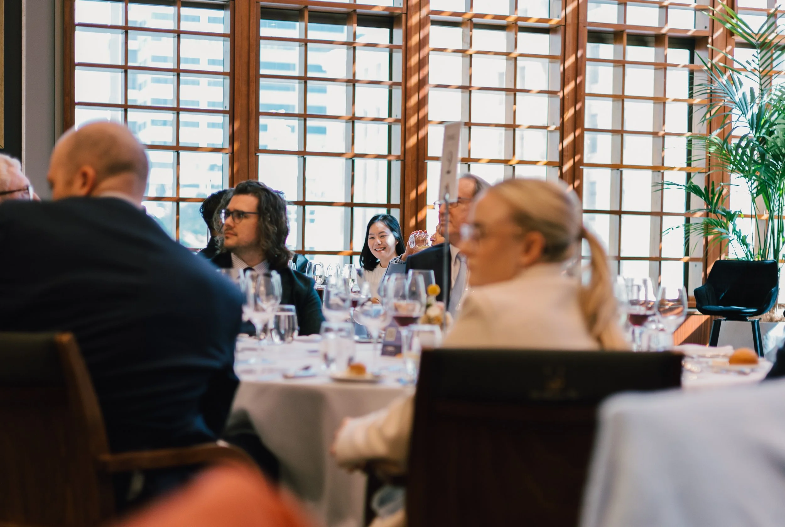 People seated at a round table during a formal event or dinner, inside a room with large grid windows and a potted plant in the background.