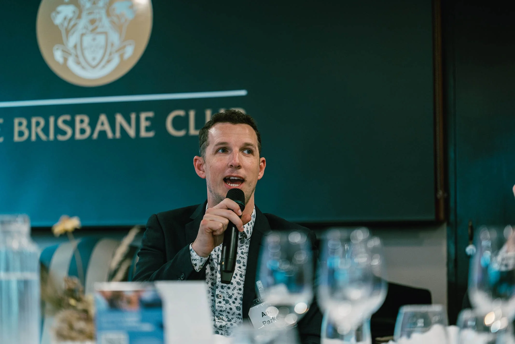 A man speaking into a microphone at a conference or event, with the Brisbane Club logo in the background.