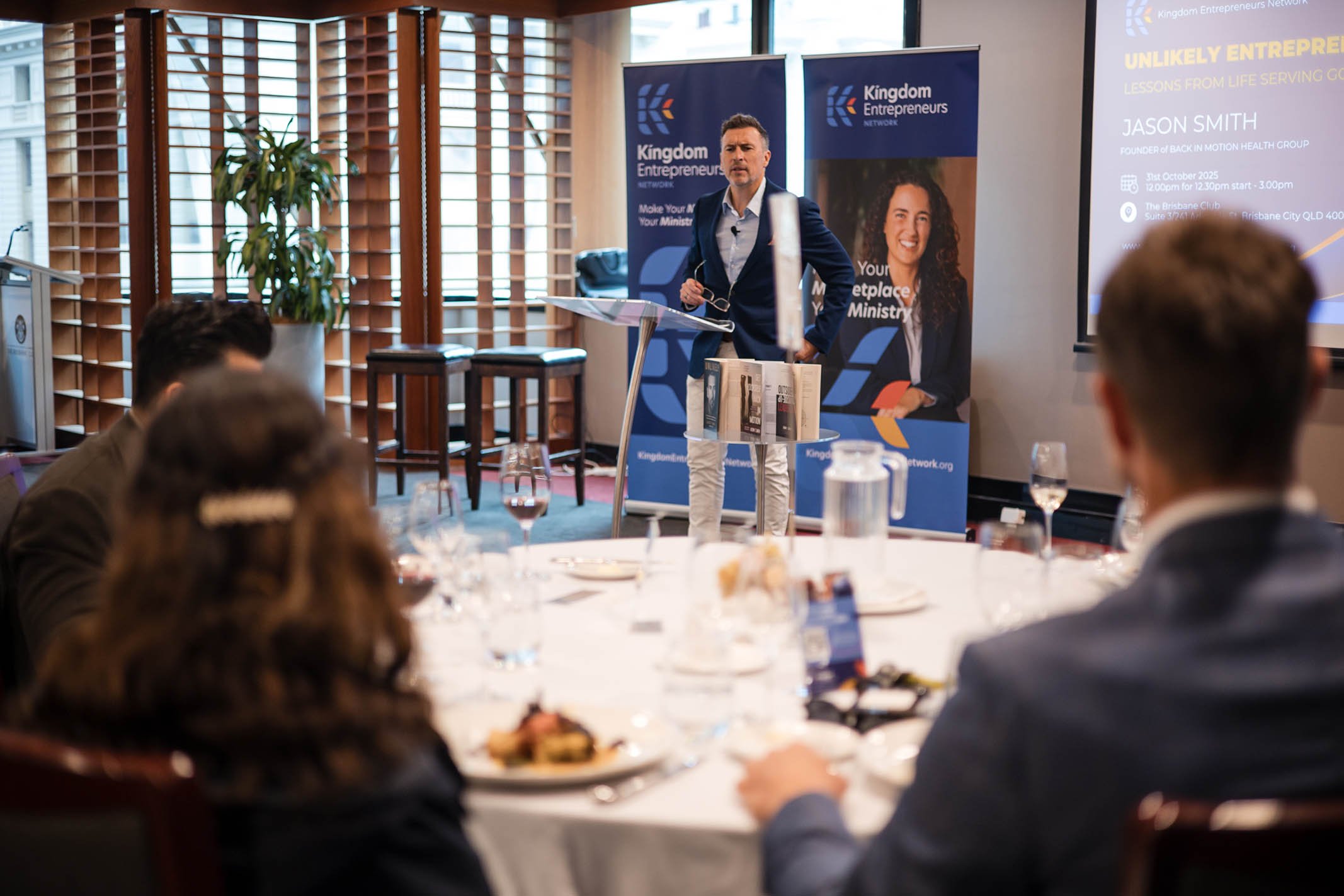 A man giving a presentation at a conference room with a screen, banners, and seated attendees.