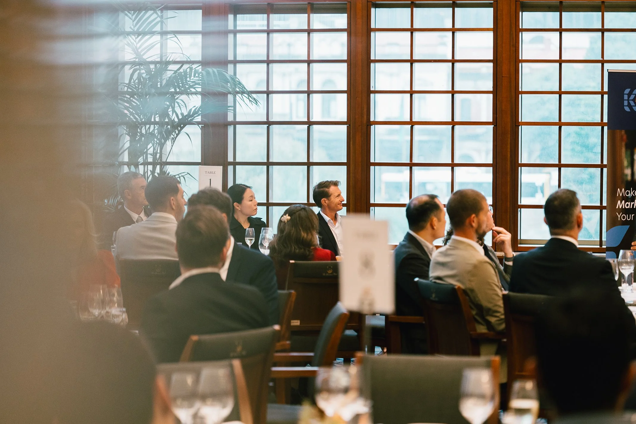 A group of business professionals seated at tables during a formal event or conference, listening attentively to a speaker or presentation, with large windows in the background allowing natural light.