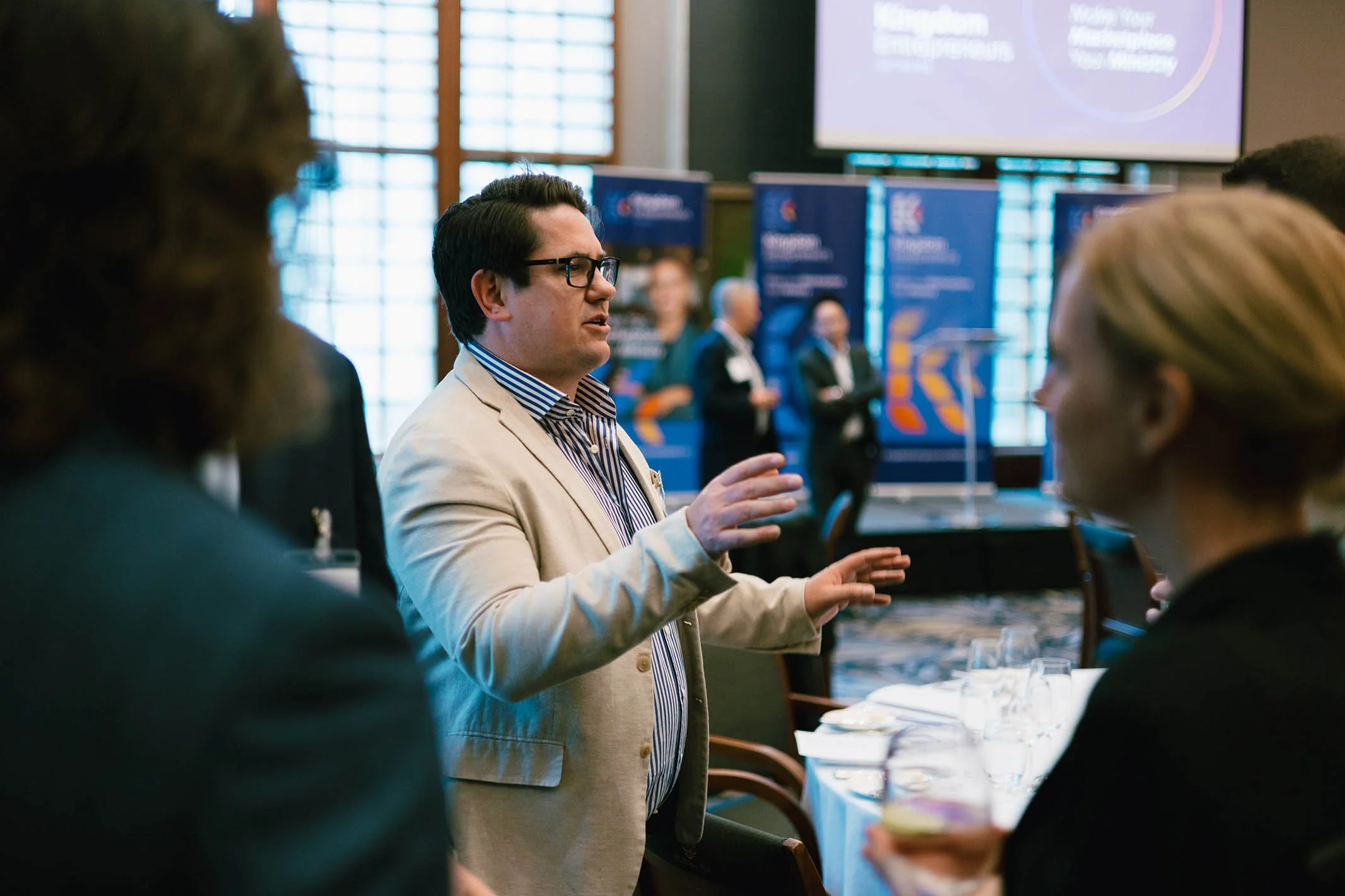 A man in a beige blazer and glasses is speaking at a professional event or conference, gesturing with his hands. Several people are gathered around him, and a large projection screen with the words 'Klamath Entrepreneur' is visible in the background.