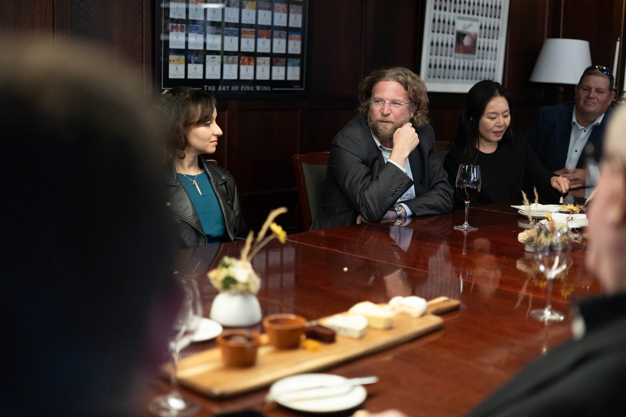 People sitting around a polished wooden table at a social gathering or meeting, with one person drinking red wine, decorated with a flower arrangement and a cheese platter, in a room with dark wood paneling and framed art on the walls.