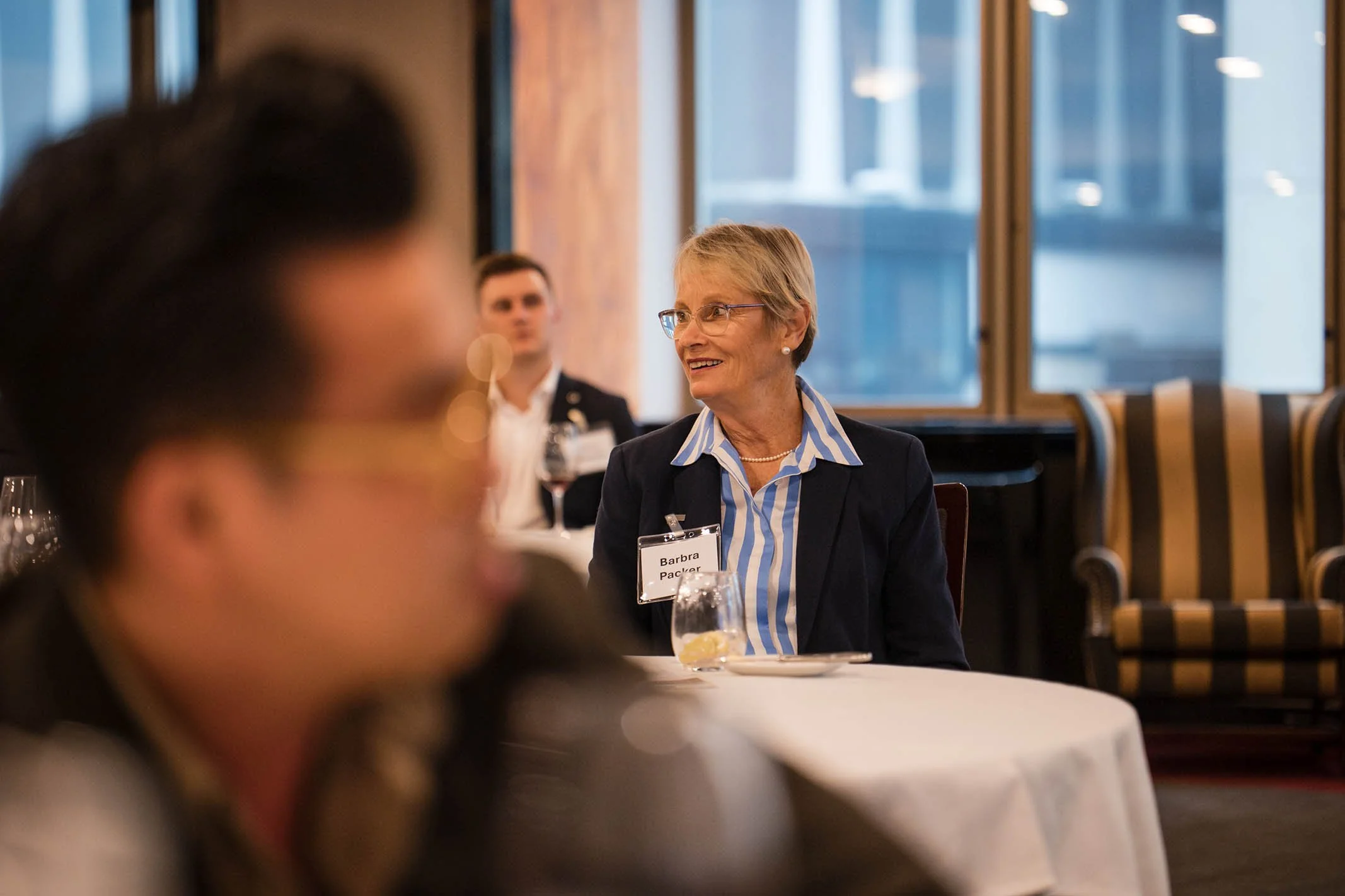 An older woman with short blonde hair and glasses, wearing a striped blue and white shirt and a dark blazer, smiling at a formal event. She has a name tag that reads "Barbra Packer". The background shows windows and people seated at tables.