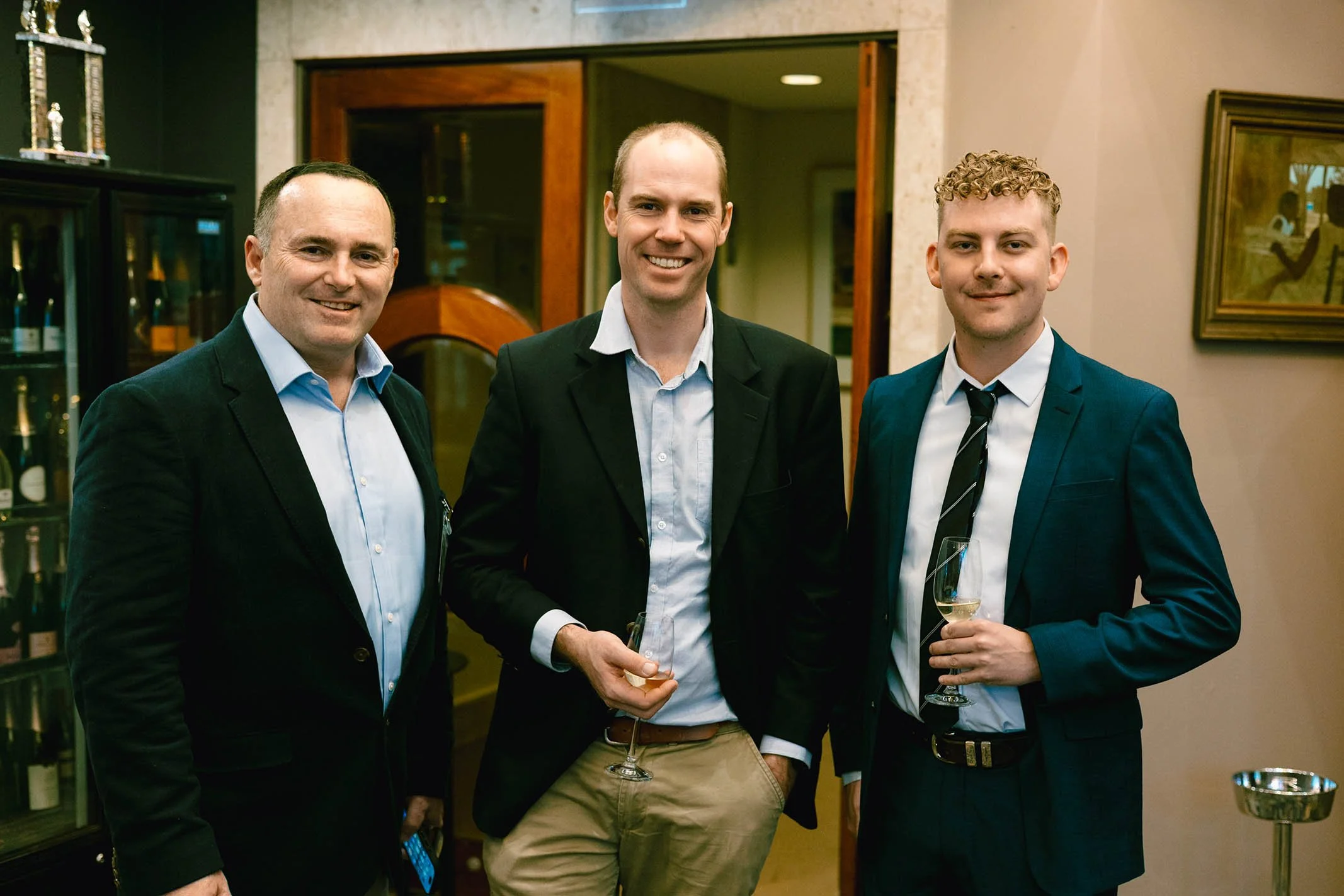 Three men in suits smiling at a social event, holding wine glasses, indoors with wine bottles in background.