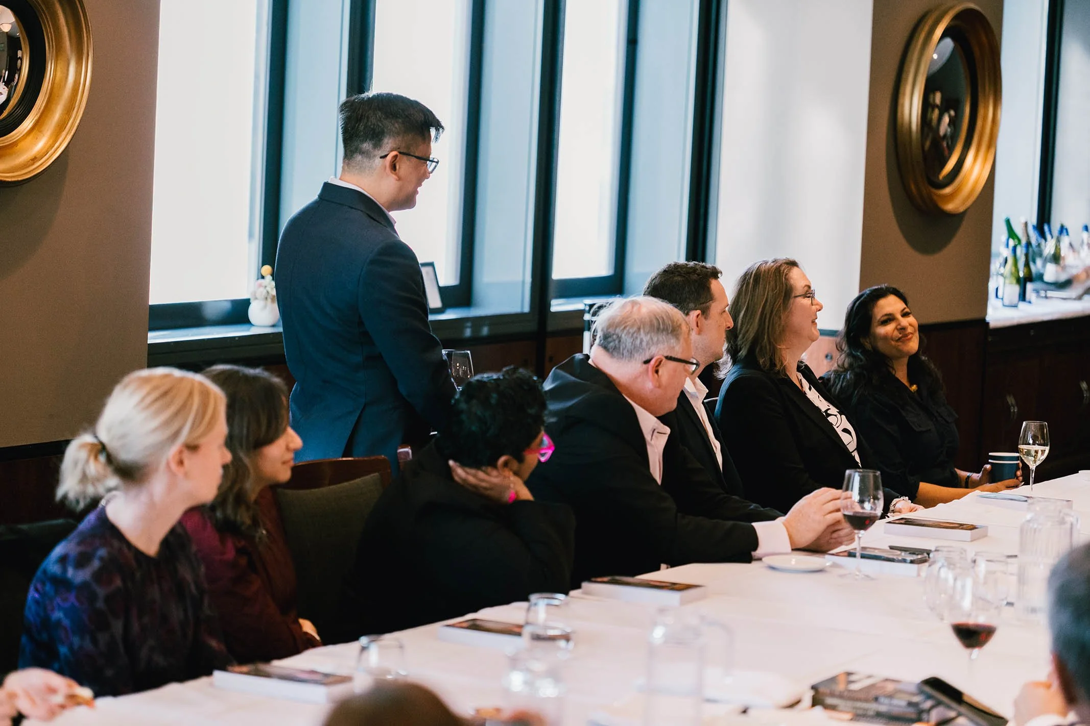 A man in a suit standing at a long table with people seated, during a formal event in a room with large windows and minimal curtains.