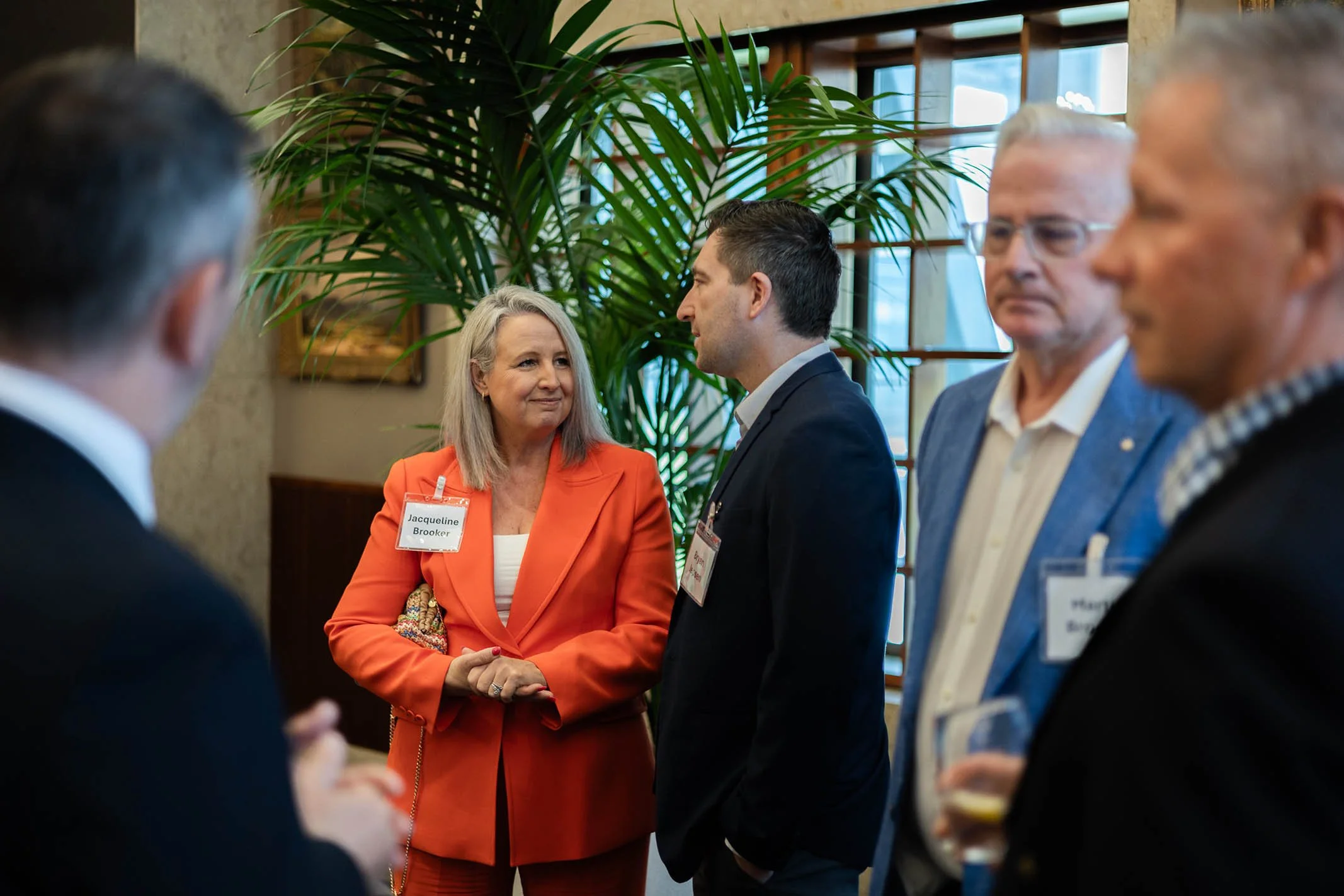 A woman in an orange blazer and gray hair talking to a man in a black suit at a professional networking event. Behind them, there are three other men, one with glasses and white hair, and another with light hair, all engaged in conversation. A large 