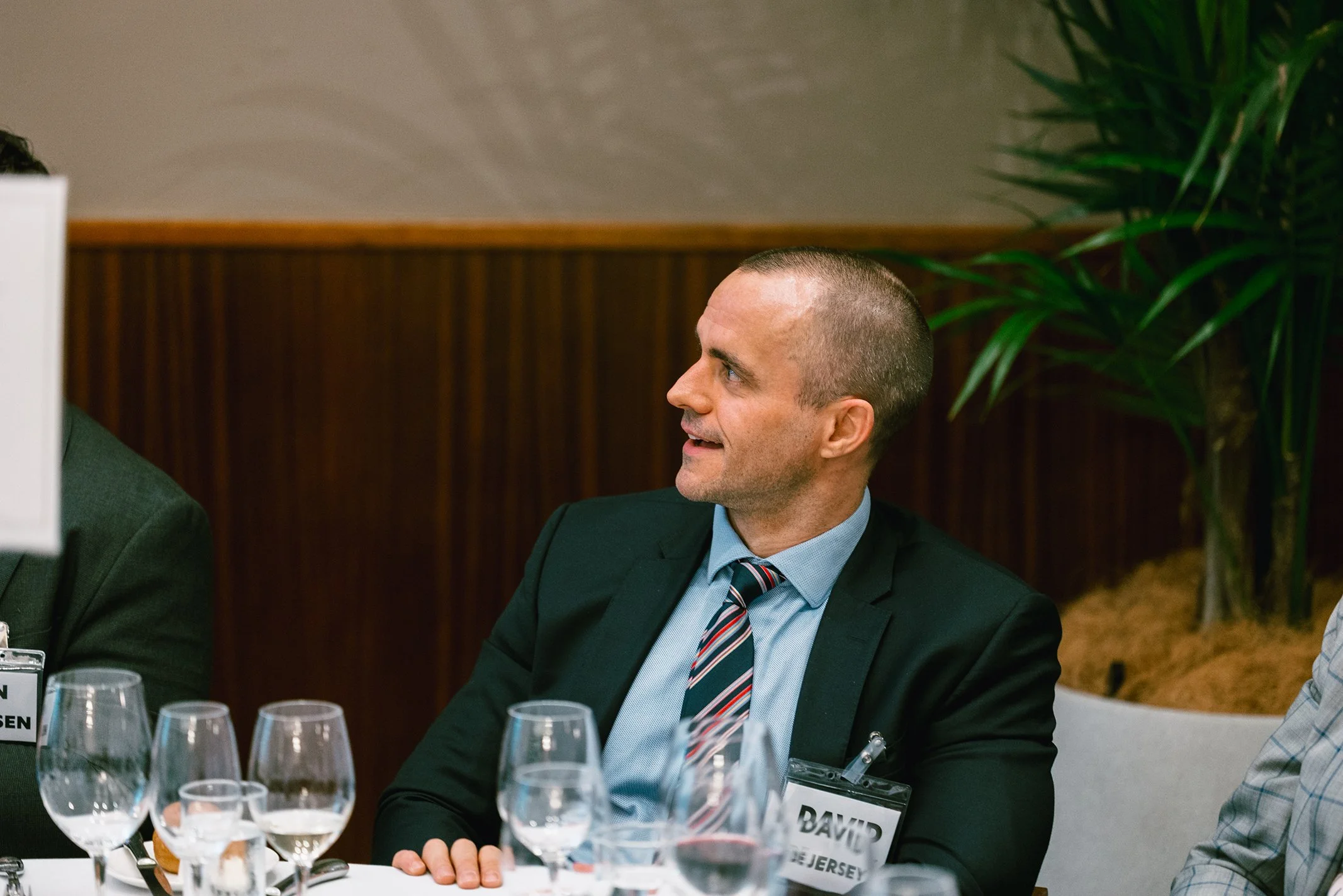 A man with short hair, wearing a suit and a striped tie, sitting at a conference table with multiple wine glasses, looking to his right.