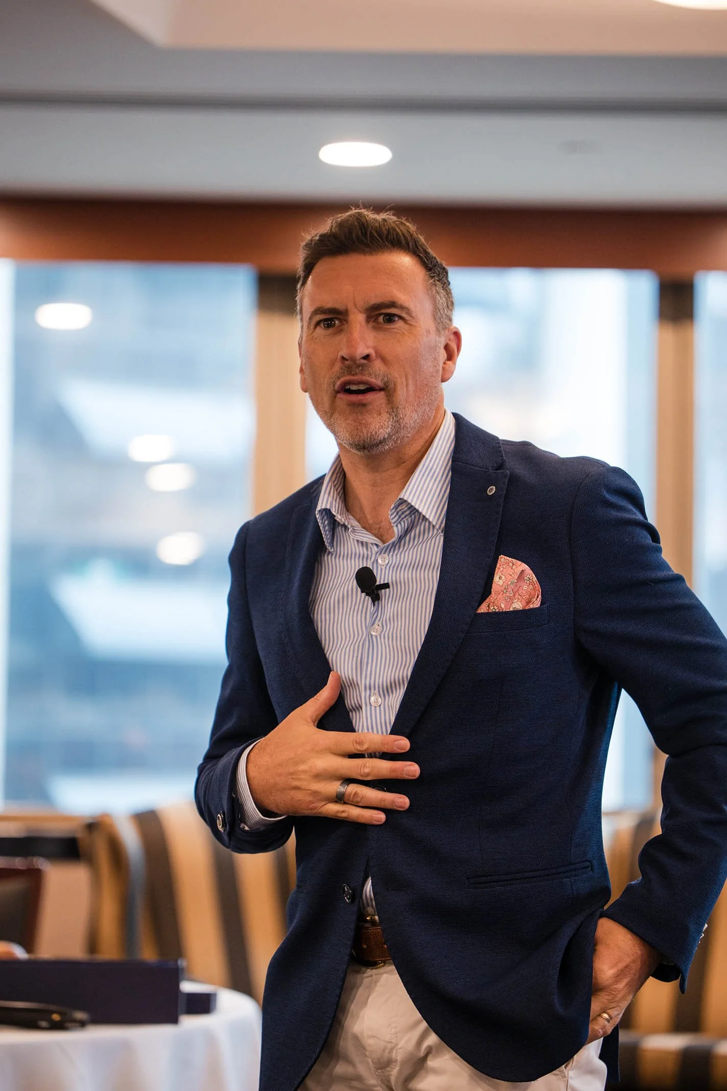 A man speaking passionately at a conference or meeting, dressed in a navy blazer, striped shirt, with a pocket square, standing in front of large windows.